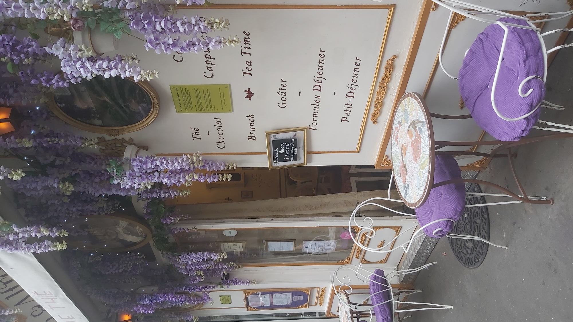 The image shows the exterior of a cafe in Paris, France, decorated with purple wisteria flowers. In the foreground, there are two white wrought-iron chairs with purple cushions and a small round mosaic-topped table. The cafe's facade is painted white with gold trim. A menu board is visible on the wall, listing various food and drink options. The cafe's entrance is partially visible, revealing a glimpse of the interior with tables and chairs. The scene suggests a charming and inviting outdoor seating area, characteristic of Parisian cafes.