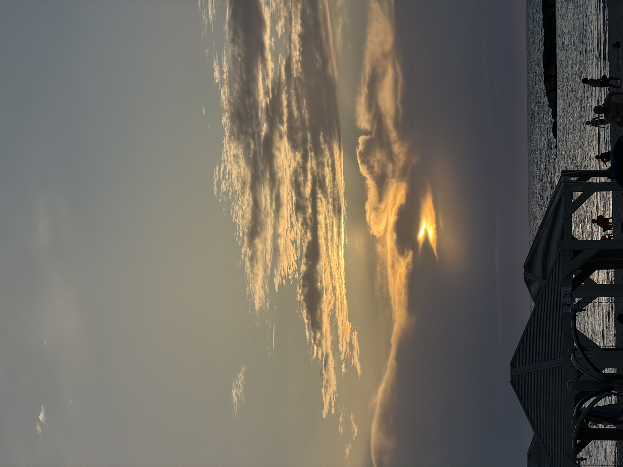 An outdoor scene captures a vibrant sunset over a body of water, likely the Mediterranean Sea in Tel-Aviv, Israel. The image is dominated by a dramatic sky filled with long, streaky clouds illuminated in brilliant golden and orange hues by the setting sun, which appears as an intense burst of light partially obscured by clouds on the right side. The sky transitions from a lighter, hazy blue-grey on the left to darker, warmer tones near the horizon.
Below the sky, the water appears dark and calm, with subtle reflections of light. Several dark silhouettes of people are visible near the water's edge on the right, some standing in the shallow water, suggesting they are enjoying the evening at the beach.
In the bottom right corner, a dark, angular structure with a slatted design, possibly a lifeguard station or a beach facility, stands against the backdrop of the sea. On this structure, two circular signs are present, though their specific symbols are indistinct due to the low light. Below these signs, a white label with the text "2 B" is visible, and further down, faint lettering like "C 3" or "G 3" can be seen, likely serving as identifiers for the beach section or facility.
The overall impression is one of peaceful relaxation at dusk, with the sun casting a warm glow over the clouds and silhouetting the figures enjoying the tranquil beach scene.