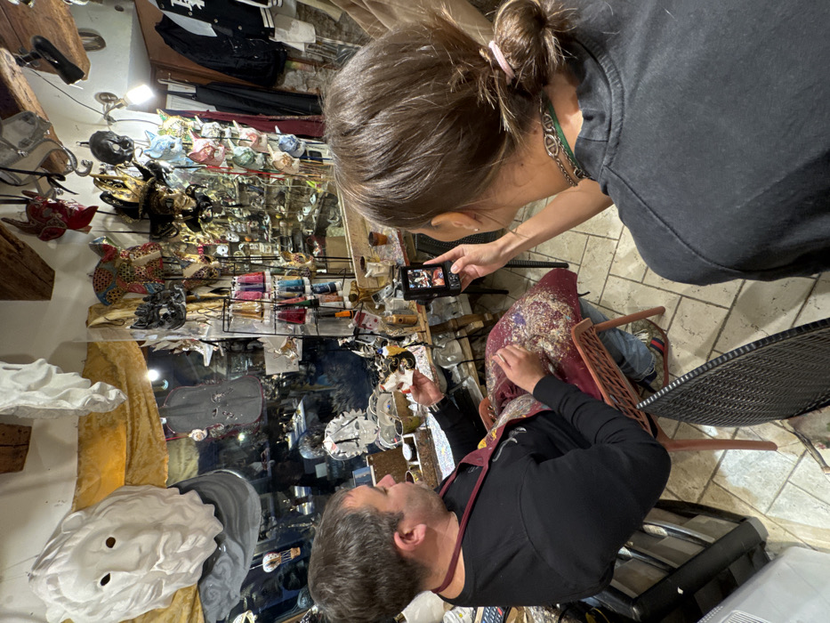 The scene depicts the interior of an artisan mask workshop located in Venice, Italy. A man with dark hair and a beard, wearing a black long-sleeved shirt and a maroon apron, is seated on the left. He is holding a small, light-colored, sculpted object, likely a mask or a mask component, in his left hand, and appears to be performing detailed work on it. His lap is covered by a patterned cushion liberally scattered with small, colorful crafting fragments. To his right, a woman, seen from behind, with brown hair tied in a bun and wearing a dark top with a necklace, holds a black smartphone horizontally, actively recording or photographing the man's activity. The workshop walls and display cases are densely populated with a diverse array of finished and unfinished Venetian masks of various styles, sizes, and colors, including a prominent large white bearded face mask on the upper left. Crafting materials and tools are visible on the workbench around the seated man. The environment features tiled flooring, exposed wooden ceiling beams, and artificial lighting.