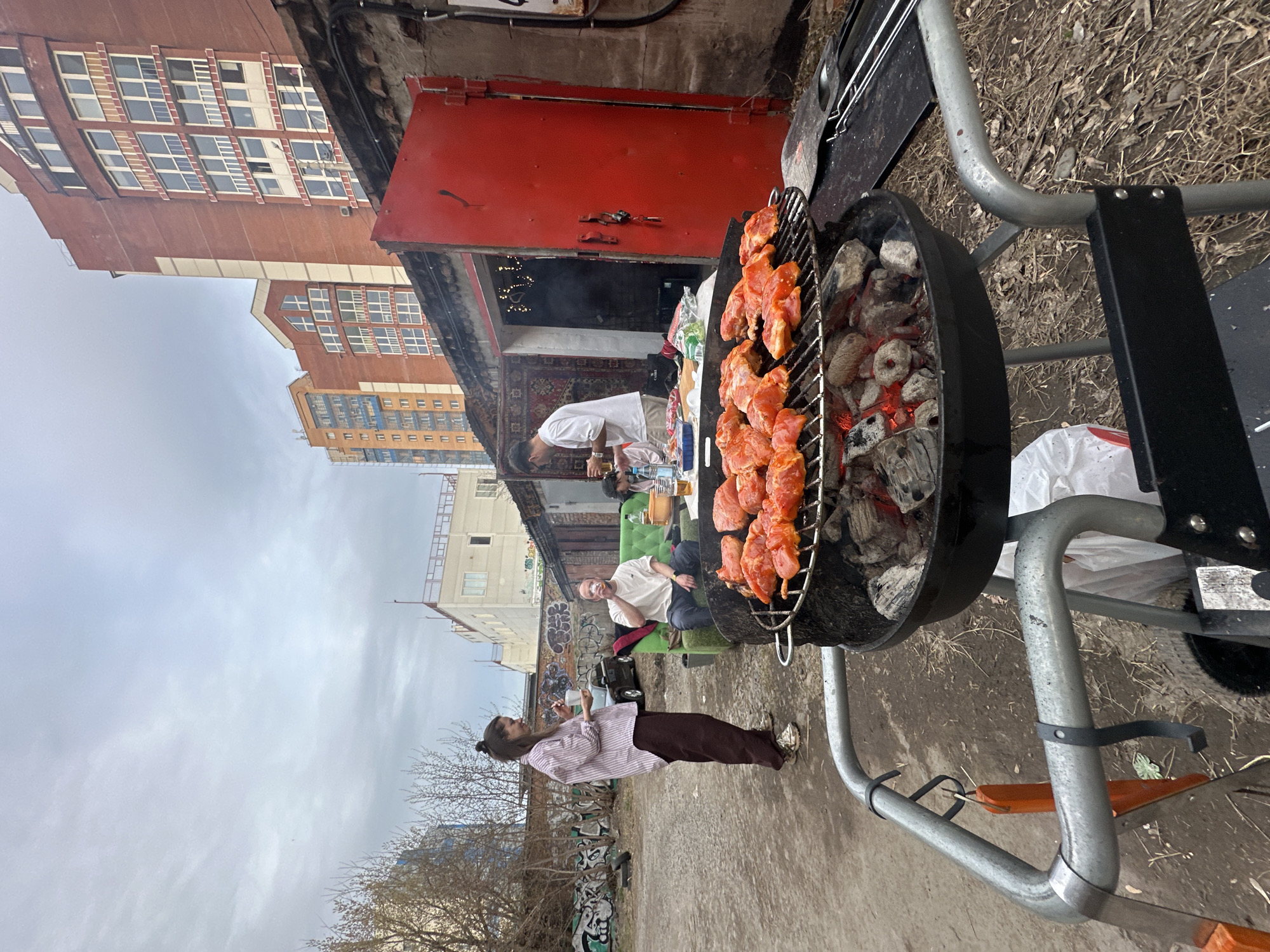 Here's a detailed description of the image:
**General Description:**
The image depicts an outdoor barbecue scene. A grill is the central focus, with raw meat cooking over hot coals. Several people are present, seemingly involved in the activity. Buildings and street are in the background, suggesting an urban location.
**People:**
* One person is standing near the grill, looking at the food being cooked.
* Another person stands inside the building behind the grill.
* Two more people are sitting on a green couch, observing or participating in the barbecue.
**Objects:**
* **Grill:** A black charcoal grill is the main object, with meat skewered and placed on the grates.
* **Coals:** Hot coals are visible below the grill, providing the heat source.
* **Buildings:** High rise buildings appear in the background.
* **Couch:** A green couch sits in the backyard of the building.
* **Red Door:** A red storage door is visible in the background.
* **Decor:** a carpet and holiday lights are visible inside of the building.
* **Drink:** A water bottle sits on the table inside.
* **City:** The setting seems to be an outside area near buildings.
**Actions and Interactions:**
* Cooking: Someone is grilling meat.
* Socializing: People are sitting, standing, and interacting, suggesting a social gathering.
**Environment:**
* **Urban Setting:** The presence of buildings and street suggests an urban area.
* **Outdoor Space:** The barbecue is taking place in an outdoor area, possibly a backyard or patio.
* **Overcast sky:** The image looks like it was taken on an overcast day.
**Location:**
I am unable to determine the exact location within the city of Krasnoyarsk, Russia, based solely on the provided image.