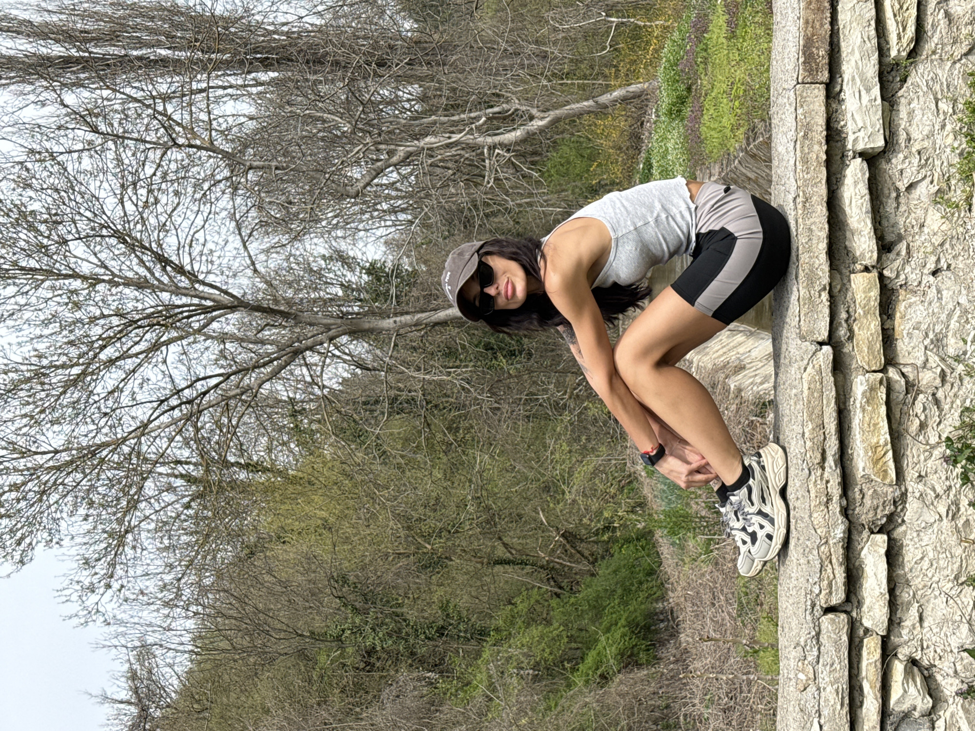 A slender woman is posed on a stone wall. She wears a gray tank top, black and gray athletic shorts, a dark baseball cap, and sunglasses. She is leaning forward, her hands holding her ankles. The background features leafless trees, other greenery, and a cloudy sky. The location is in the city of Pleven, Bulgaria.