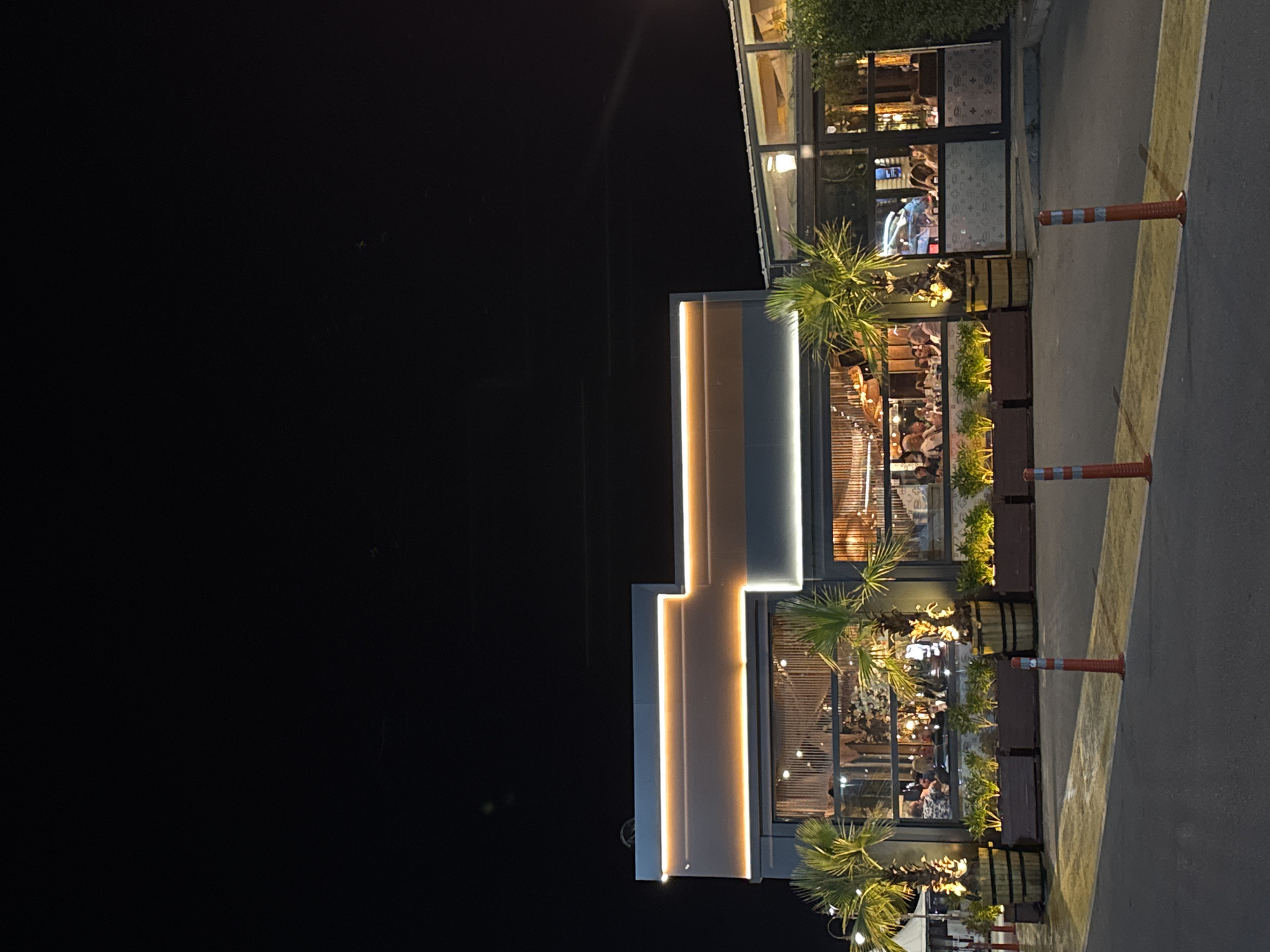 At an unknown urban location, a modern, multi-story commercial building, likely a restaurant or cafe, is illuminated at night. The structure features a dark grey facade with prominent horizontal strip lighting in a warm orange hue along its roofline and mid-sections. Large glass windows on the ground floor reveal a bustling interior where numerous individuals are seated at tables, engaged in dining or social interaction.
In front of the main building, several palm trees, illuminated by upward-pointing lights, are housed in large wooden barrel planters. Between these, elongated rectangular planters contain smaller green foliage, also lit with warm light. To the right of the main building, an attached glass-enclosed structure, possibly an additional dining area or conservatory, is visible, with its interior lights on and people inside. A dense green hedge borders the far right of the property.
The immediate foreground consists of a dark asphalt surface with distinct yellow painted lines or markings. Three orange and white striped traffic cones (bollards) are positioned on this surface, two on the right and one on the left, casting small shadows. The sky above is completely dark, indicative of nighttime.