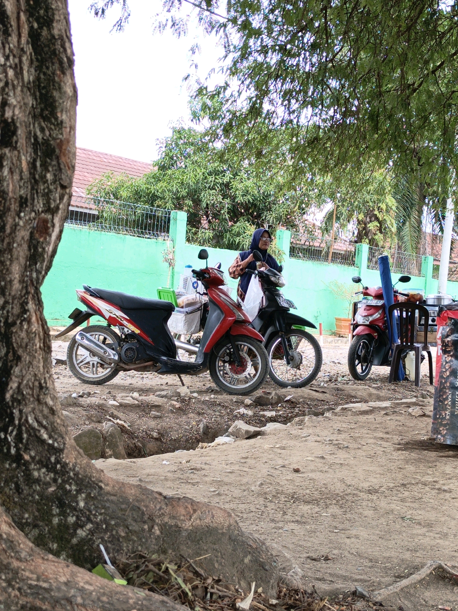 In Gedung Nyapah, Indonesia, an outdoor scene depicts several motorcycles parked on a dirt lot. A large tree trunk dominates the left foreground. Behind the motorcycles, a vibrant green fence runs horizontally, with a red-tiled roof structure and trees visible beyond it.
A woman wearing a hijab and glasses stands between two motorcycles, holding what appears to be a plastic bag. The motorcycles are a red and black scooter on the left, and a black scooter in the middle. To the right, another red motorcycle is parked next to a brown plastic chair. On the chair, a large rolled-up blue mat is propped up. A metal cart with what looks like a food steamer and other items is positioned behind the chair and the red motorcycle. The ground is rough and uneven, with scattered rocks and debris. The overall impression is a simple, possibly commercial or residential, outdoor area.