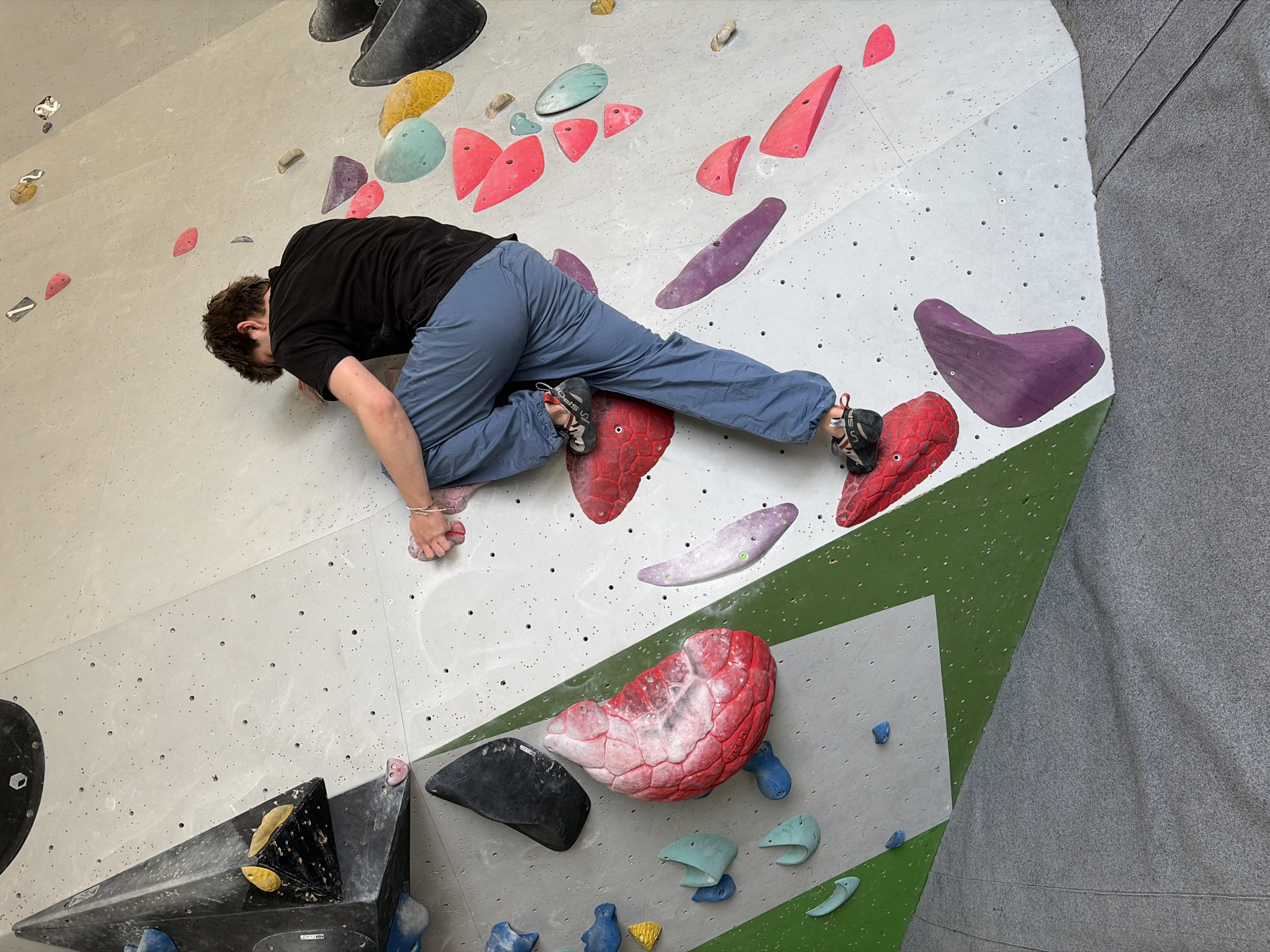 A person wearing a black t-shirt and blue pants is rock climbing indoors on a white and green wall with multicolored holds. The person is positioned horizontally, with their hands gripping a small, light-colored hold and their feet placed on two large, red holds. Various other climbing holds of different colors and shapes are visible on the wall. The location is likely a climbing gym in Ipswich, United Kingdom.