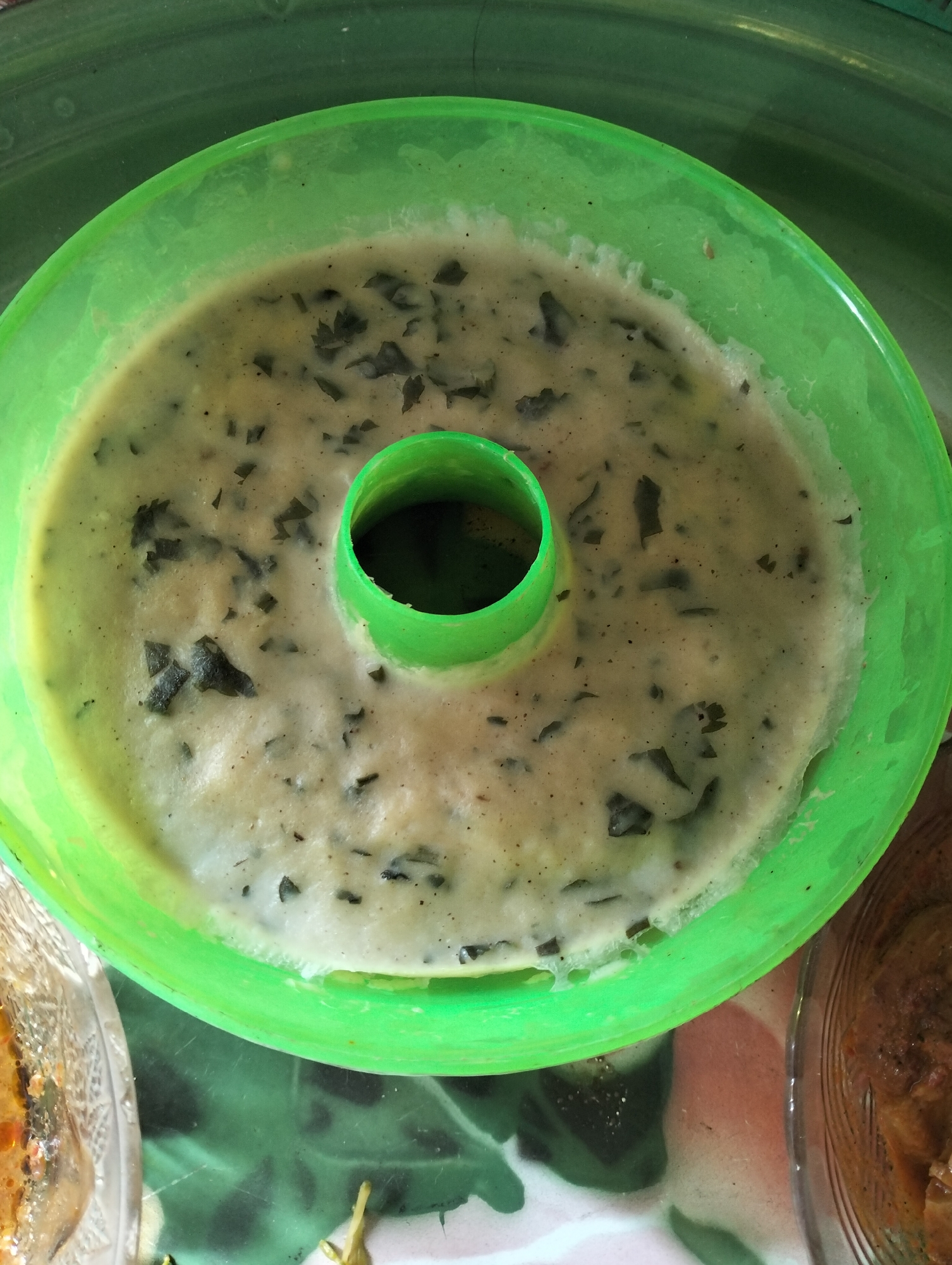 An overhead, close-up shot shows a bright green, ring-shaped baking pan filled with a light-colored batter. The batter contains small, dark green flecks, likely herbs or vegetables. A darker green, cylindrical mold rises from the center of the pan, creating the ring shape. The pan is sitting on a green surface with a patterned texture. To the left and right of the pan, parts of clear glass dishes containing food are visible. The food in the dish to the right appears to be a brownish, stew-like dish. The image is taken from a high angle, emphasizing the contents of the baking pan. There are no people visible in the image. This image was likely taken in a kitchen in Mandala Jaya, Indonesia, judging by the provided context.