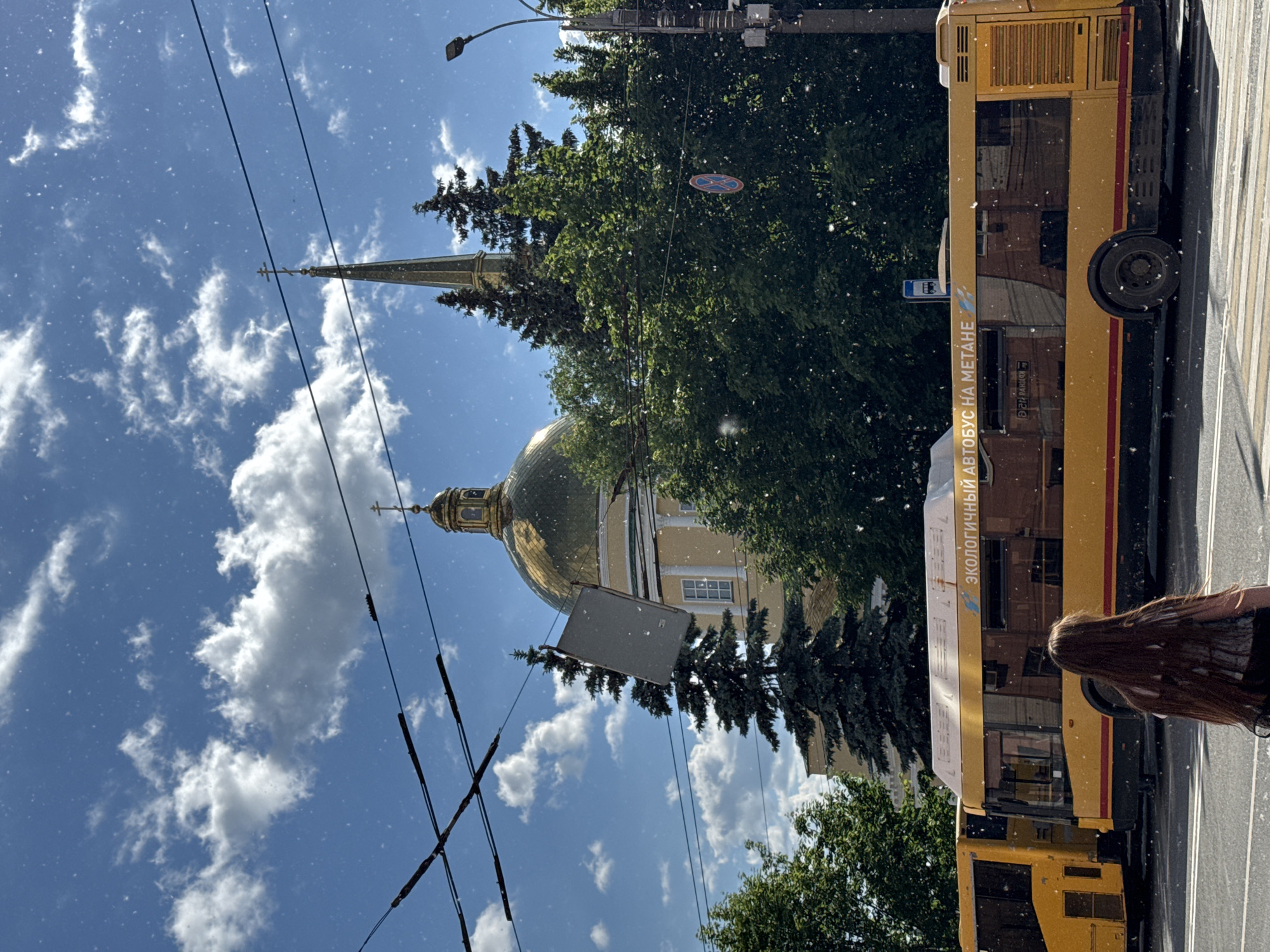 This vertical full shot depicts a bright, sunny daytime scene on a city street in Izhevsk, Russia. The image is filled with numerous small white specks, resembling airborne pollen or fluff, against the clear blue sky and darker elements.
In the mid-ground, a prominent yellow bus occupies the right side of the frame, appearing to be at a stop or moving slowly. The bus features the Russian text "ЭКОЛОГИЧНЫЙ АВТОБУС НА МЕТАНЕ" (ECOLOGICAL BUS ON METHANE) printed on its side. A person with long, dark hair is visible from behind in the lower right corner, seemingly observing the street or waiting near the bus.
Behind the bus and to the left, a dense canopy of green trees fills much of the center, partially obscuring a majestic building. This building is a church or cathedral, characterized by a shining golden dome and a tall, slender spire topped with a cross, typical of Russian Orthodox architecture. A rectangular grey object hangs from wires in front of the church.
Overhead, multiple black power lines stretch across the blue sky, which is dotted with scattered white clouds. A street light is visible in the upper right. Among the trees, a round red and blue road sign can be discerned. The overall impression is a bustling urban environment on a warm, clear day, possibly during late spring or summer given the abundant green foliage and visible pollen.