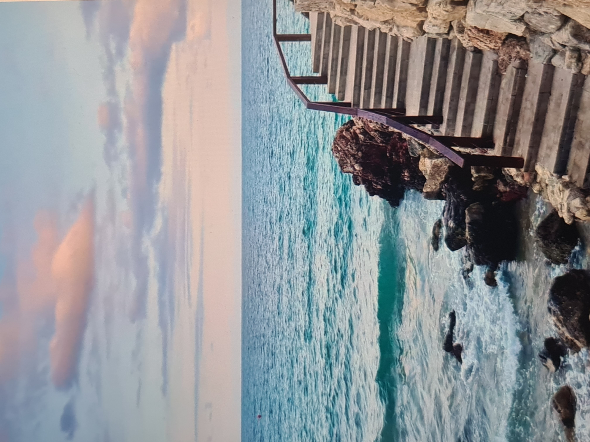 The image depicts a serene coastal landscape featuring a set of stairs descending towards the ocean. The scene is oriented vertically, showing the horizon line running horizontally across the middle.
On the right side of the image, a robust stone or concrete staircase, approximately one person wide, is carved into or built along a rocky cliff face. A dark-colored metal handrail, possibly made of wood or iron, follows the descent of the stairs, providing support. Large, rough rocks form the immediate edge of the land, some partially submerged in the water.
The ocean dominates the central and lower portions of the image, displaying clear, turquoise-blue waters with visible ripples and small waves, particularly closer to the shore where they break against the rocks, creating white foam. The water extends to the horizon, meeting a soft, expansive sky.
The sky, occupying the left side and upper portion, is painted with gentle hues of light blue, fading into soft pinks and oranges towards the top left, indicative of either sunrise or sunset. Wispy, diffused clouds with warm tones are scattered across the sky, adding to the tranquil atmosphere.
No people or identifiable events are taking place, suggesting a moment of natural beauty. There is no visible text. The overall impression is one of calm and natural splendor, with the man-made stairs providing access to the pristine waters below.