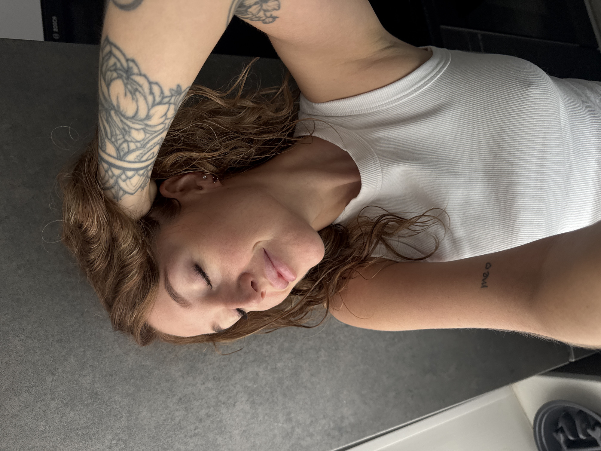 A close-up, high-angle shot captures a young woman with reddish-brown, curly hair, her eyes closed and a gentle smile on her face. She is wearing a white ribbed tank top and has her right arm raised, hand resting on her head, revealing tattoos on her arm. Her left forearm also shows a small tattoo with the word "me" and a heart. She appears to be indoors, with a textured grey wall behind her and a glimpse of a black appliance with a "BOSCH" logo in the background. The lighting suggests natural light from the side. No specific location in Chaikovsky, Russia can be determined from the visual information.