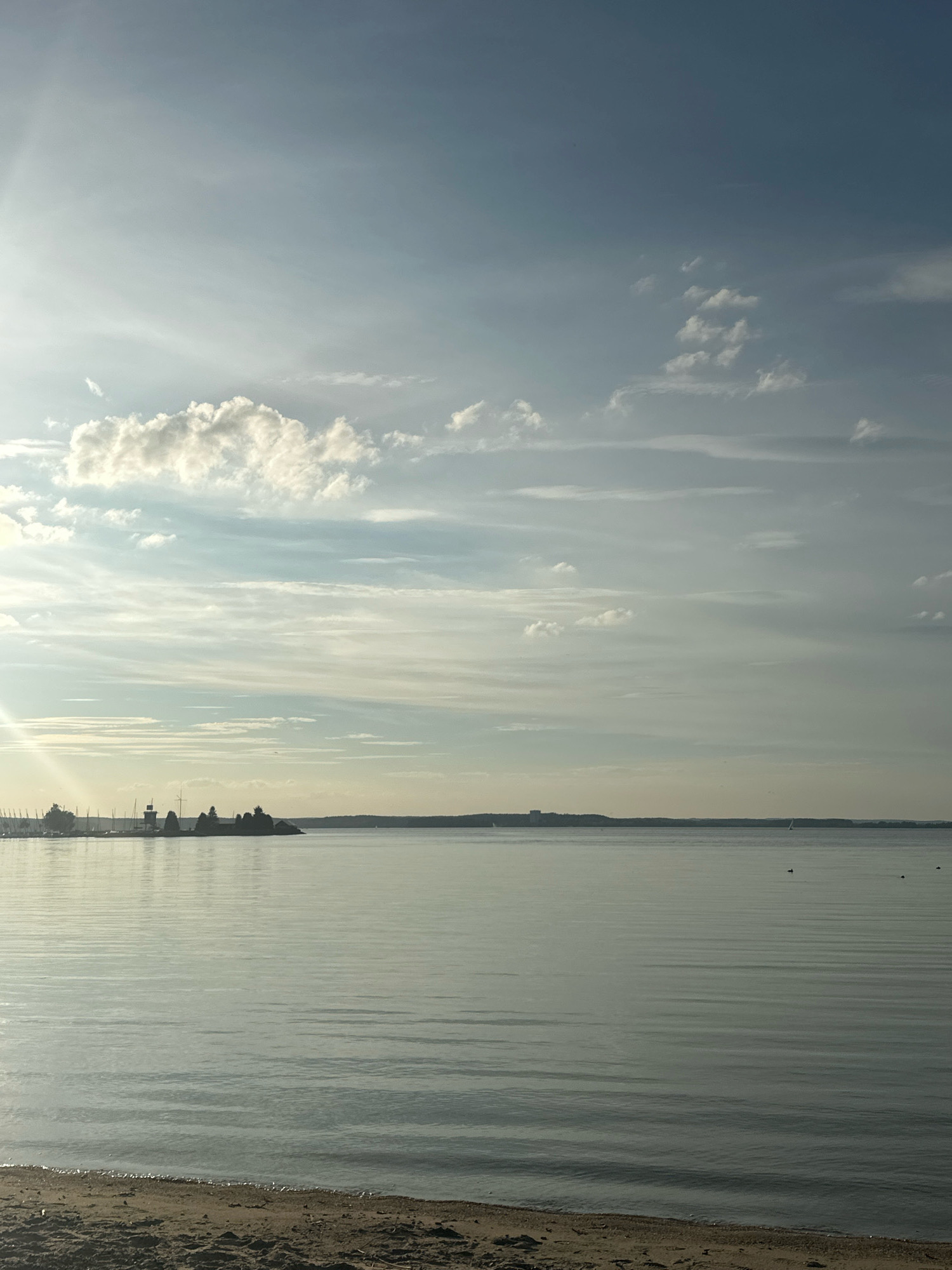 The image depicts a serene waterscape with a sandy shore in the foreground and a calm body of water stretching towards a distant, tree-lined horizon. The sky is a blend of pale blue and white clouds, with the sun casting a gentle glow on the scene, creating a bright, slightly hazy atmosphere. On the left side of the image, a marina with several sailboats and a lighthouse is visible. The water's surface is smooth, with subtle ripples reflecting the sky. The overall impression is one of tranquility and natural beauty. The location is likely in the vicinity of Ждановіцкі сельскі Савет, Belarus, given the presence of a marina and a sizable body of water, which could be a lake or a wide river. There are no people or animals clearly visible in the image.