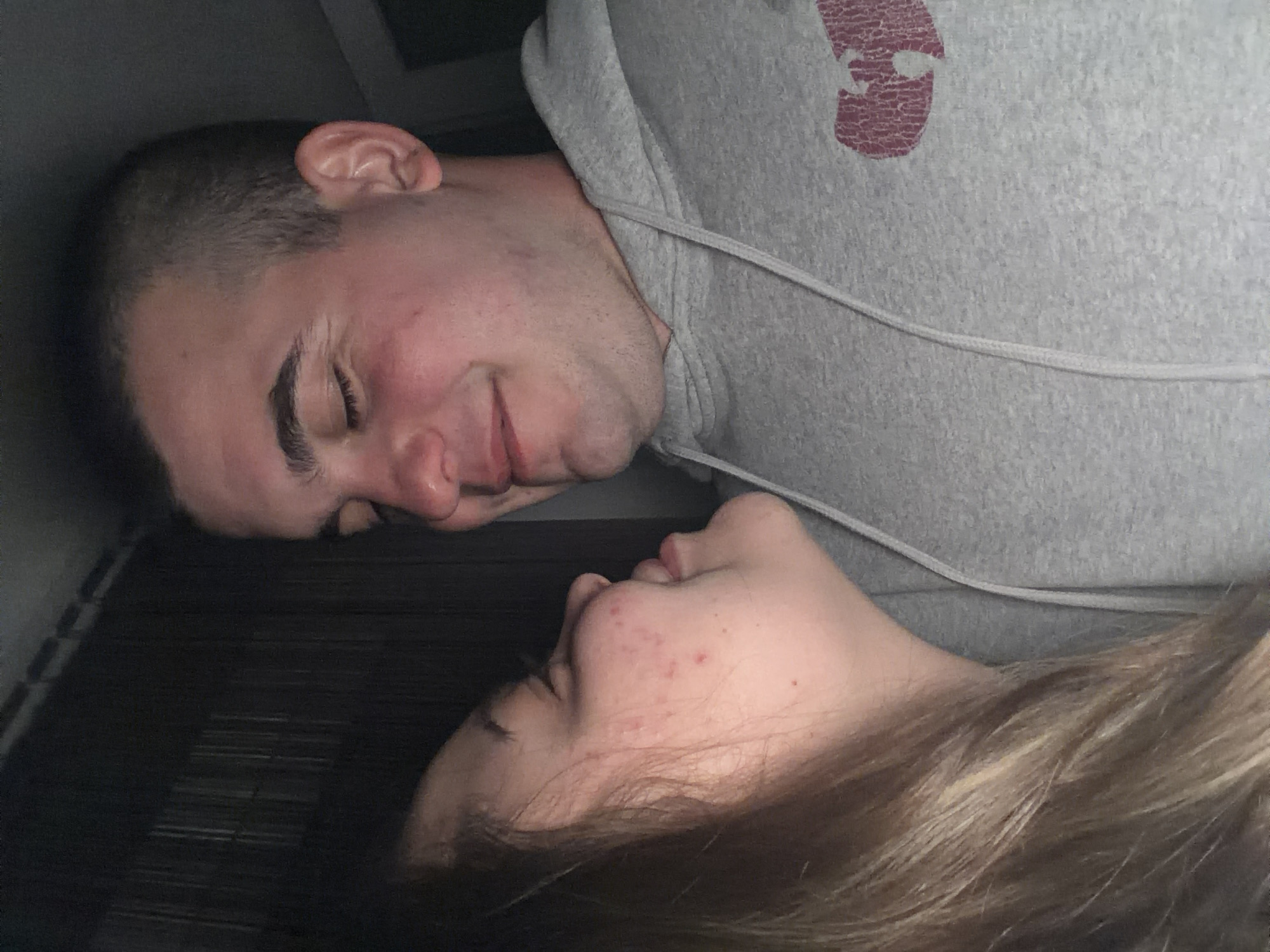 An intimate overhead shot depicts two individuals, a male and a female, lying horizontally and facing each other in a dimly lit indoor setting. The image is rotated ninety degrees counter-clockwise from a standard upright view.
The male, positioned on the left, has short, dark hair and appears to be resting peacefully, with his eyes closed or looking downwards, a soft, content smile gracing his lips. He is wearing a grey hooded sweatshirt with a visible white drawstring. On the right side of his chest, there's a faded, reddish-pink graphic featuring a distressed texture, which could be a stylized letter or an abstract logo.
The female, on the right, has lighter, possibly blonde or light brown, wavy hair. Her face shows some acne, and her eyes are open, gazing towards the male, suggesting a moment of connection or admiration. She is also wearing a grey garment, likely a part of the same hooded sweatshirt or similar attire.
Both subjects are lying on a dark, textured surface that features horizontal lines, possibly indicating a bed or a couch with a ribbed pattern. The close proximity and their expressions convey a tender, affectionate, and comfortable moment between the two. The low lighting suggests the scene takes place in the evening or at night.
While the location context is Varna, Bulgaria, there are no specific visual cues within the image itself to confirm this geographical setting. No distinct text is clearly legible on the clothing or in the background.