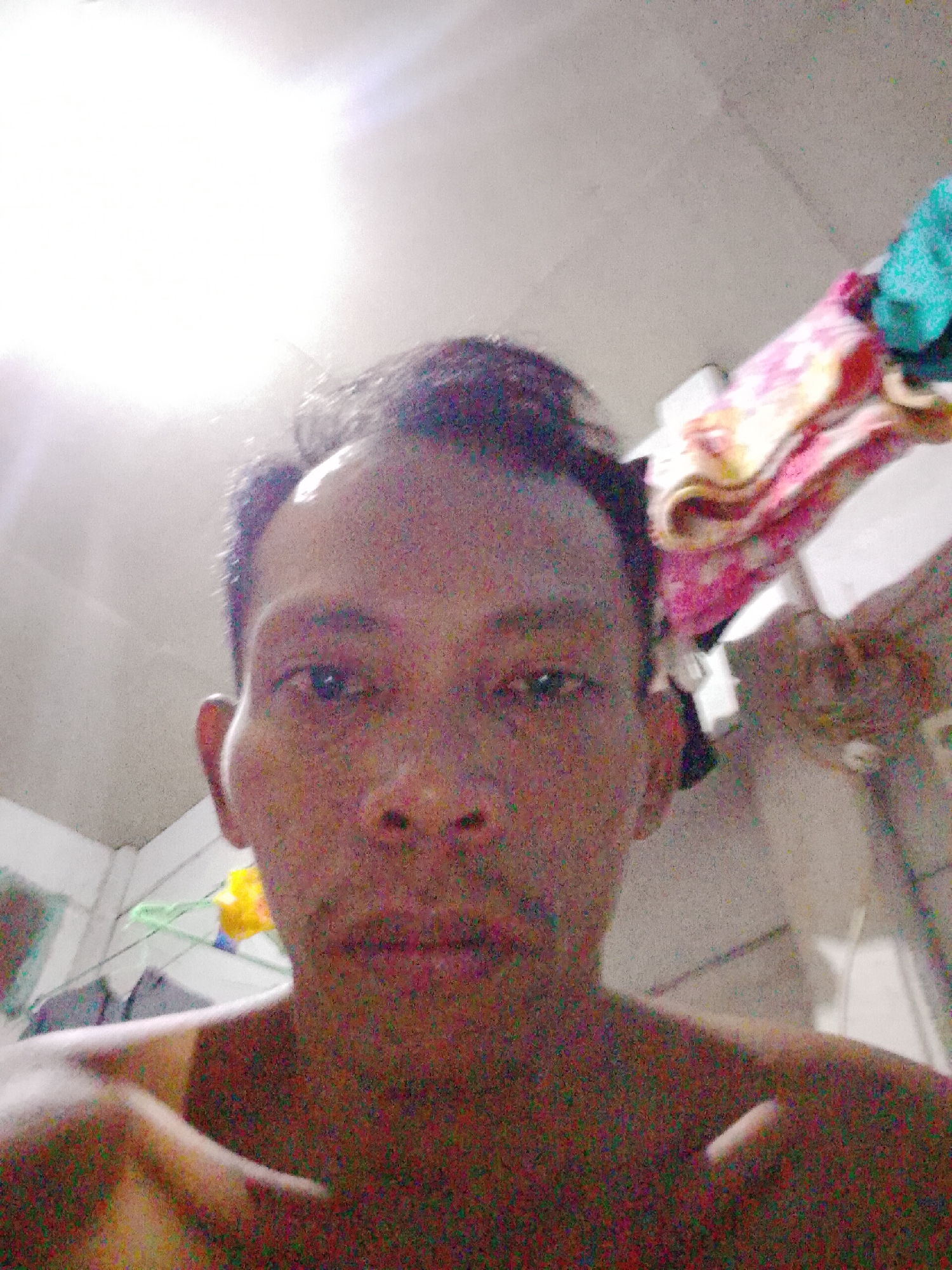 A close-up selfie photograph features an adult male with short, dark hair and a thin build. The background is slightly blurry, revealing a white ceiling, what appear to be articles of clothing, and possibly a fan. The image exhibits some noise, and the lighting is bright, resulting in a slightly washed-out appearance. The location is Bukit Ketok, Indonesia.