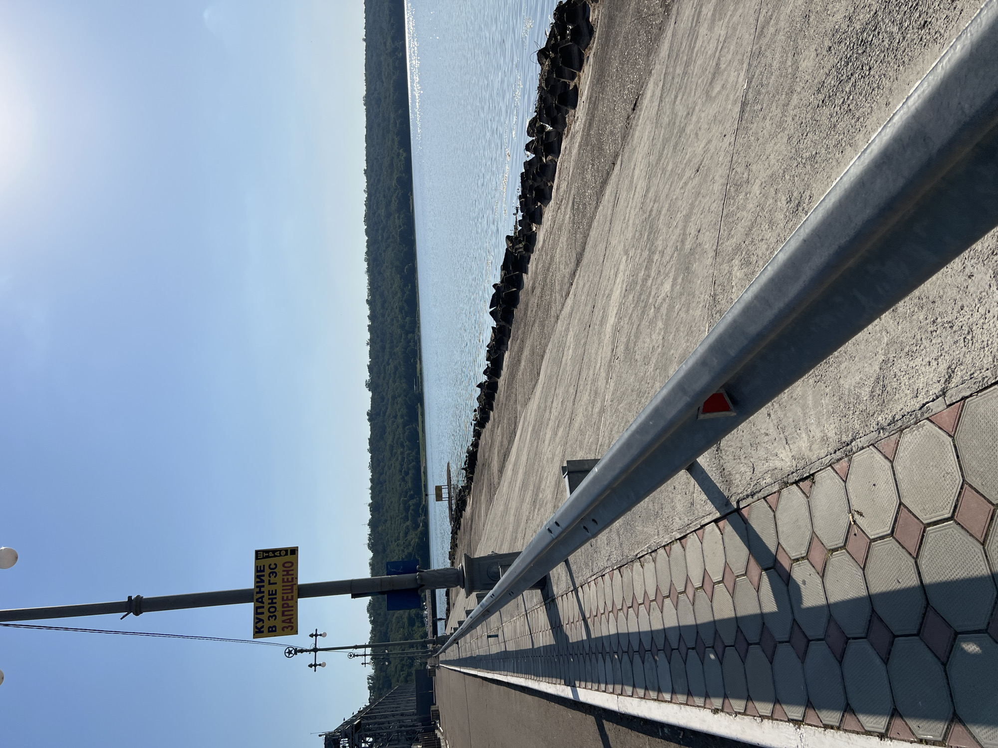 The image depicts a waterfront scene, possibly along a river or reservoir, with a clear blue sky. On the left, there is a paved walkway with a distinctive hexagonal tile pattern, bordered by a concrete embankment. A metal guardrail runs along the edge of the walkway. In the background, a large industrial structure, likely a bridge or dam, is visible. The water extends to the horizon, where a tree-covered shoreline forms a natural boundary. The sun is shining, casting long shadows from the guardrail and lampposts. A yellow sign on a lamppost reads "КУПАНИЕ В ЗОНЕ ГЭС ЗАПРЕЩЕНО", which translates to "SWIMMING IN THE HPP ZONE IS PROHIBITED". The location is identified as Korzhevo, Moldova.