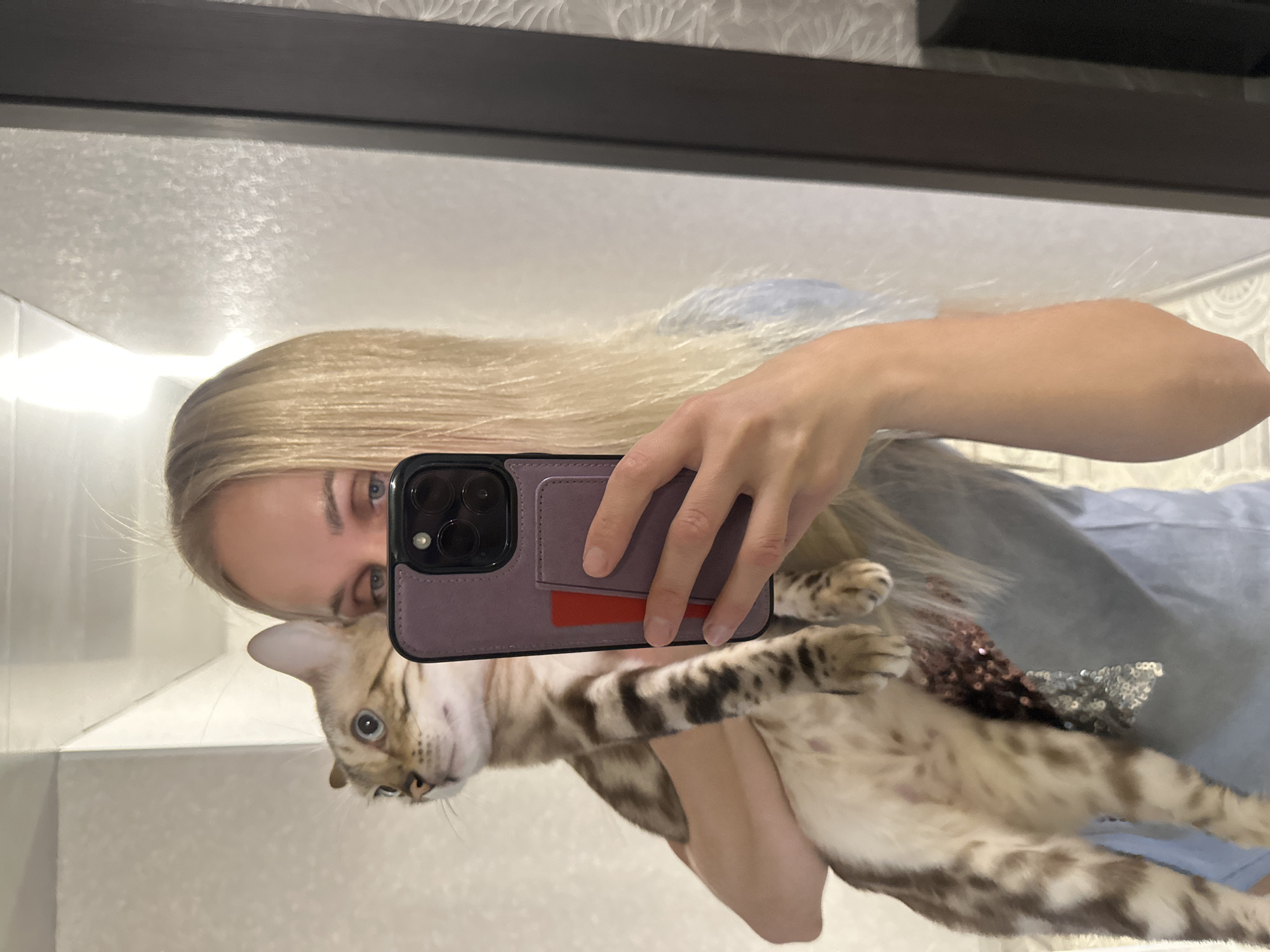 A woman with blonde hair is taking a selfie in a bathroom, holding a cat. She is wearing a light blue shirt. The cat is light brown with dark spots and is looking to the left. The bathroom has white tiled walls and a mirror. The reflection shows the woman holding the cat and her phone. The image does not contain any information that indicates the location in the city of Energetik, Russia.