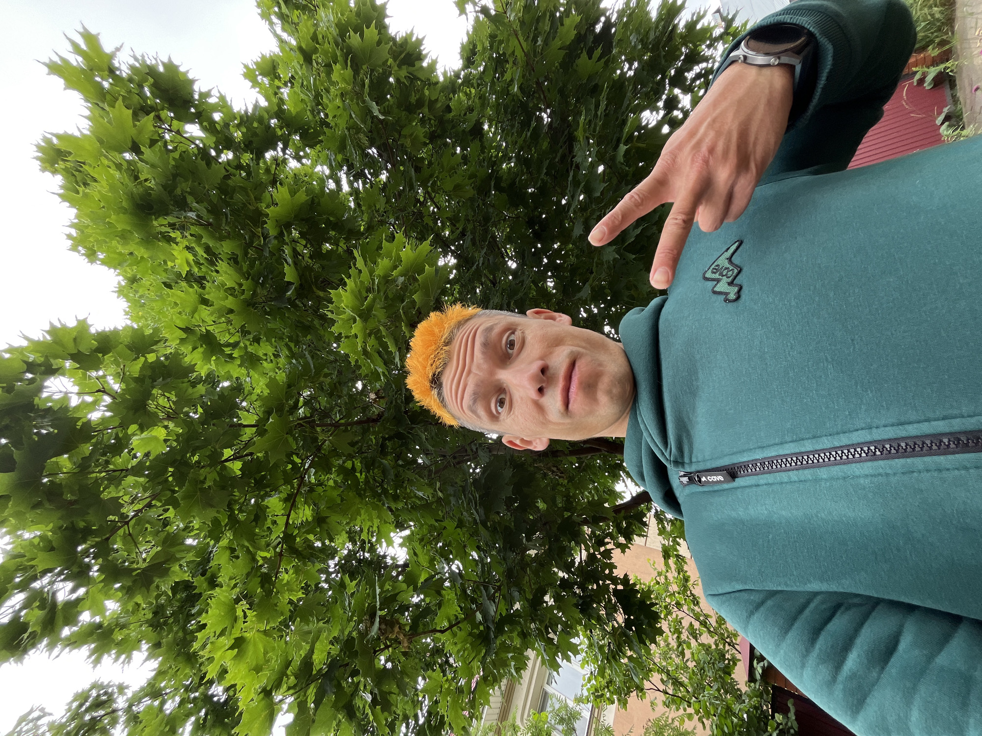 A man with bright orange hair, wearing a teal hooded sweatshirt with an asymmetrical zipper and a logo on the left chest, poses for a selfie outdoors. He is making a peace sign with his right hand. Behind him, a large green tree with full foliage dominates the frame, with hints of a building and a red fence in the background. The scene appears to be in a residential area of Chaikovsky, Russia, given the presence of a building and fence typical of such environments.