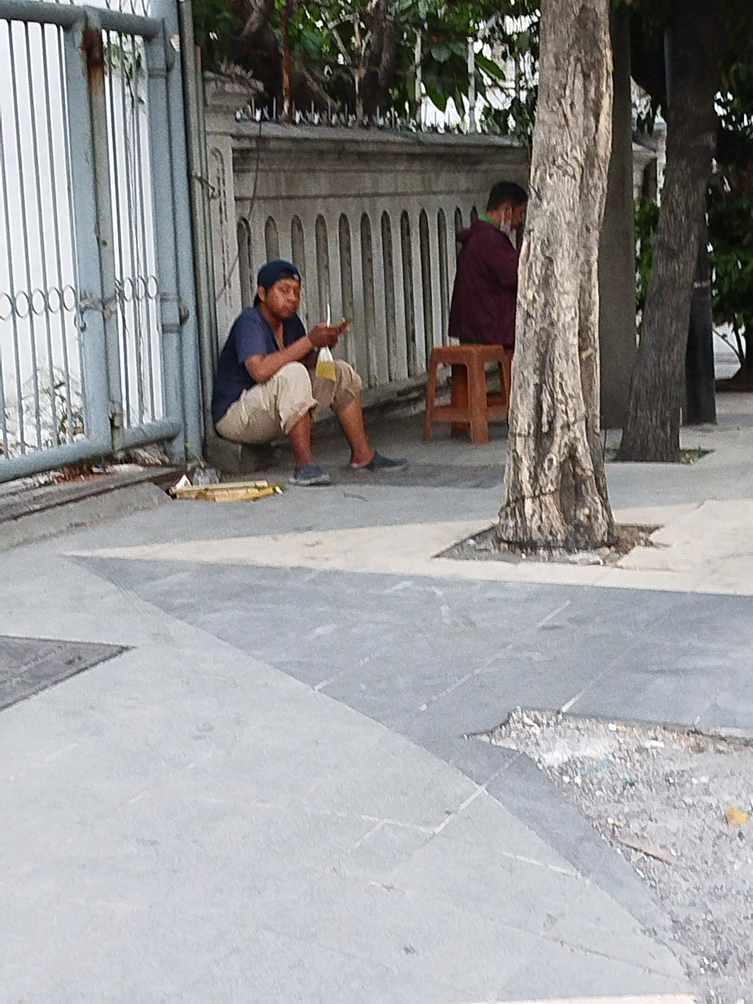 The image depicts an outdoor scene on a sidewalk in Surabaya, Indonesia, near a gated entrance. In the foreground, a person wearing a blue cap, a dark blue t-shirt, and light-colored shorts is seated on the ground. They are holding a small object, possibly food or a cigarette, in their right hand and a plastic bag with a yellow liquid in their left hand. The person appears to be eating or drinking. Beside them on the ground are some discarded items, including what looks like bamboo sticks and a plastic bottle.
In the mid-ground, another person is visible sitting on a small orange stool. This individual is wearing a dark maroon jacket and a face mask. They are partially obscured by a tree trunk. Behind the people is a concrete wall with decorative vertical elements and a metal fence. The environment suggests a somewhat urban setting, with a paved sidewalk and a glimpse of greenery and trees. The overall impression is a snapshot of everyday life on a street in Surabaya.