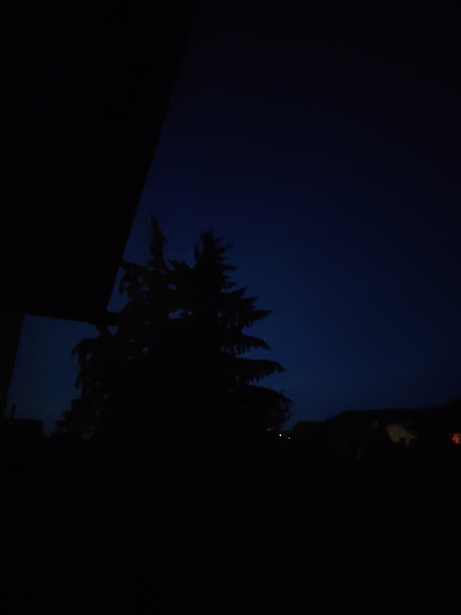 This is a low-light photograph taken at dusk or dawn in the city of Lozen, Bulgaria. The foreground is dominated by a dark, silhouetted structure on the left, possibly a balcony or overhang. To the right, the silhouette of a coniferous tree stands against a deep blue sky, which has a slight gradient, appearing darker towards the top. In the distance, the faint silhouettes of buildings and houses are visible along a horizon line, with a few small, indistinct lights suggesting artificial illumination. The overall impression is of a quiet, dimly lit evening or early morning scene in a residential or semi-rural area of Lozen.