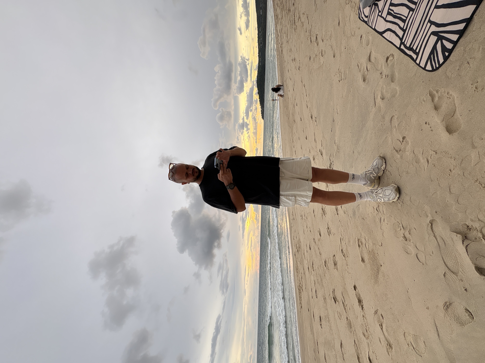 A man is standing on a sandy beach, holding a phone to take a photo of himself, or perhaps to film. He is wearing a black t-shirt and white shorts. He has glasses on and appears to have a goatee. His shoes are white athletic shoes with grey accents, and he is wearing white socks. Next to him, further up the beach, a black and white patterned blanket is visible. The man is looking down at his phone with a neutral expression.
The scene is a beach with a shoreline visible in the background. The sun is low in the sky, creating a warm glow on the horizon, suggesting it is either sunrise or sunset. The sky is partly cloudy with dramatic grey clouds scattered amongst lighter ones. The sand is light tan and covered in footprints. The ocean waves are gentle. There is a person in the distance on the beach, appearing to be a woman wearing white. The location is Choeng Thale, Thailand. There is no visible text in the image.