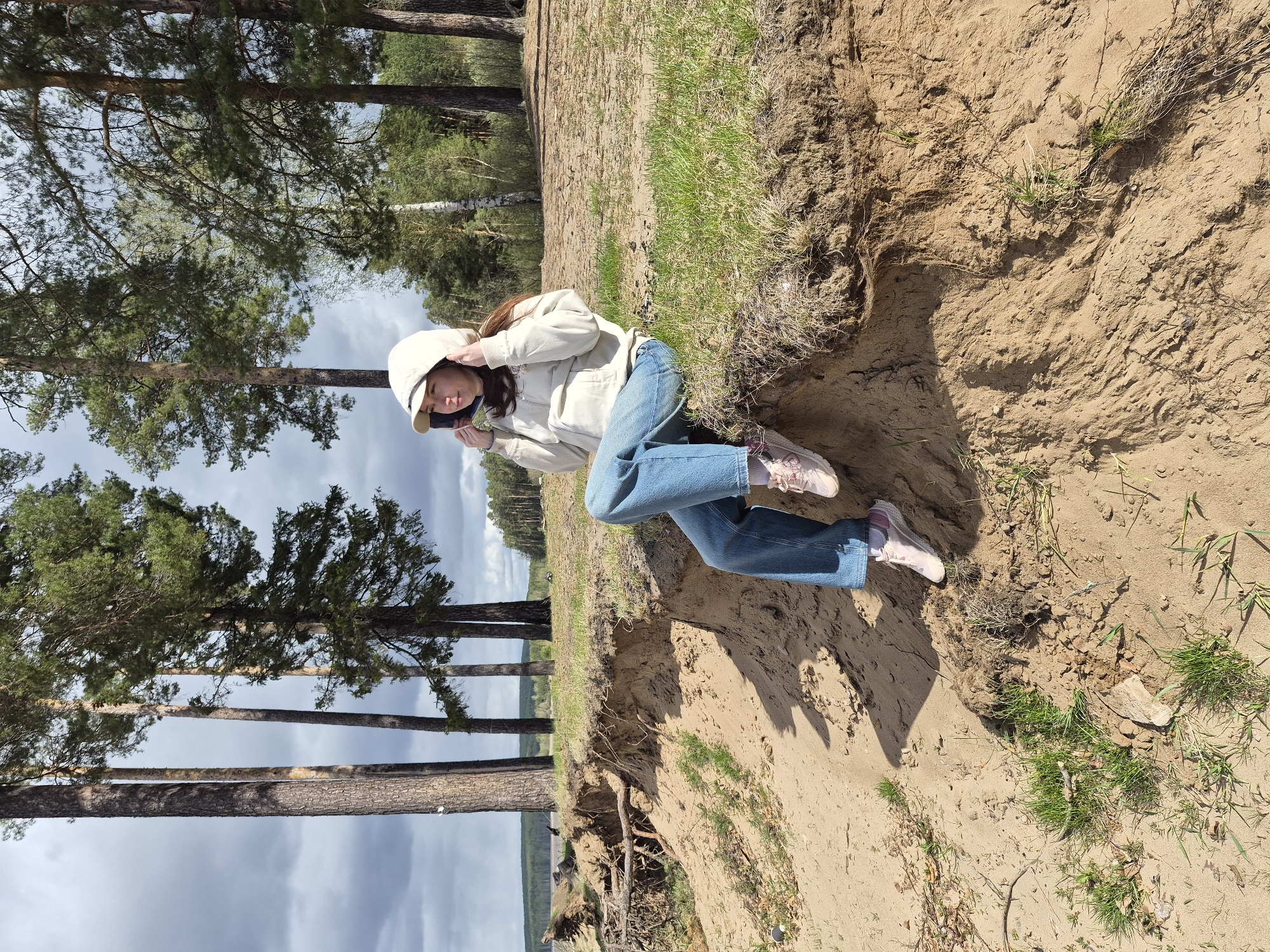 A woman wearing a beige jacket, blue jeans, and a beige cap is sitting on a sandy embankment with sparse grass. She appears to be holding something in her hands near her face. The background features a forest of tall pine trees under a cloudy sky. The location is likely a coastal area near Chaikovsky, Russia, given the presence of pine trees, sandy soil, and a body of water visible in the distance.