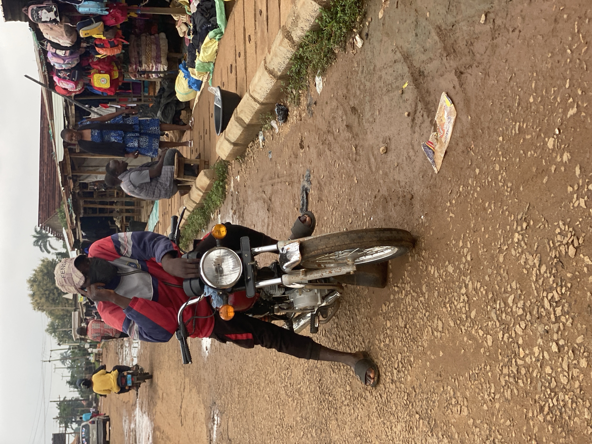A man in a red and blue jacket, black pants, and sandals is sitting on a motorcycle in Kafanchan B, Nigeria. He is holding a phone to his ear with his left hand and wearing a cap with the word "FENCH" visible. The motorcycle has a round headlight and amber turn signals. The ground is a dirt road with gravel and some patches of mud. To the left, people are standing near a market stall with bags and clothes displayed. Another motorcycle is driving away from the camera in the distance. A strip of packaging is on the ground in front of the motorcycle. The weather appears overcast. A concrete curb runs along the edge of the road with grass and weeds growing.