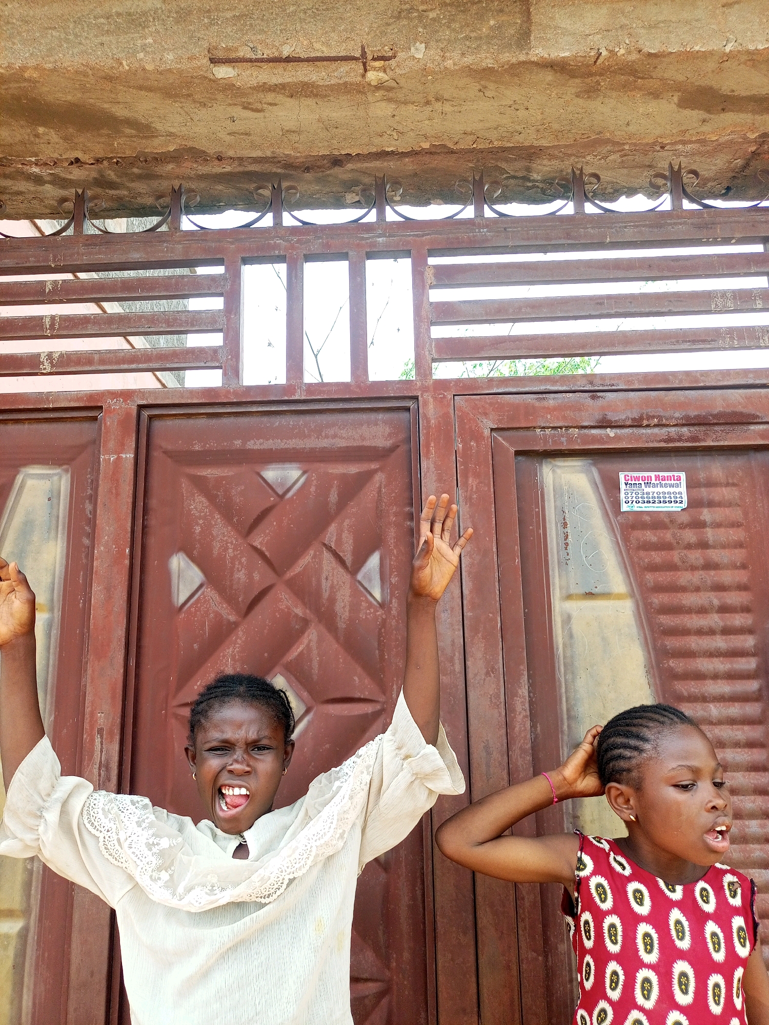 Two young girls, one with braided hair and a cream-colored top, the other with braided hair and a red patterned dress, stand in front of a brown metal gate in Tempure, Nigeria. The girl on the left is shouting with her mouth open and arms raised. The girl on the right has her hand on the back of her head and is looking to the right with her mouth open. The gate has a decorative metalwork top. A sign with text and phone numbers is visible on the right side of the gate. The concrete structure above the gate appears weathered.