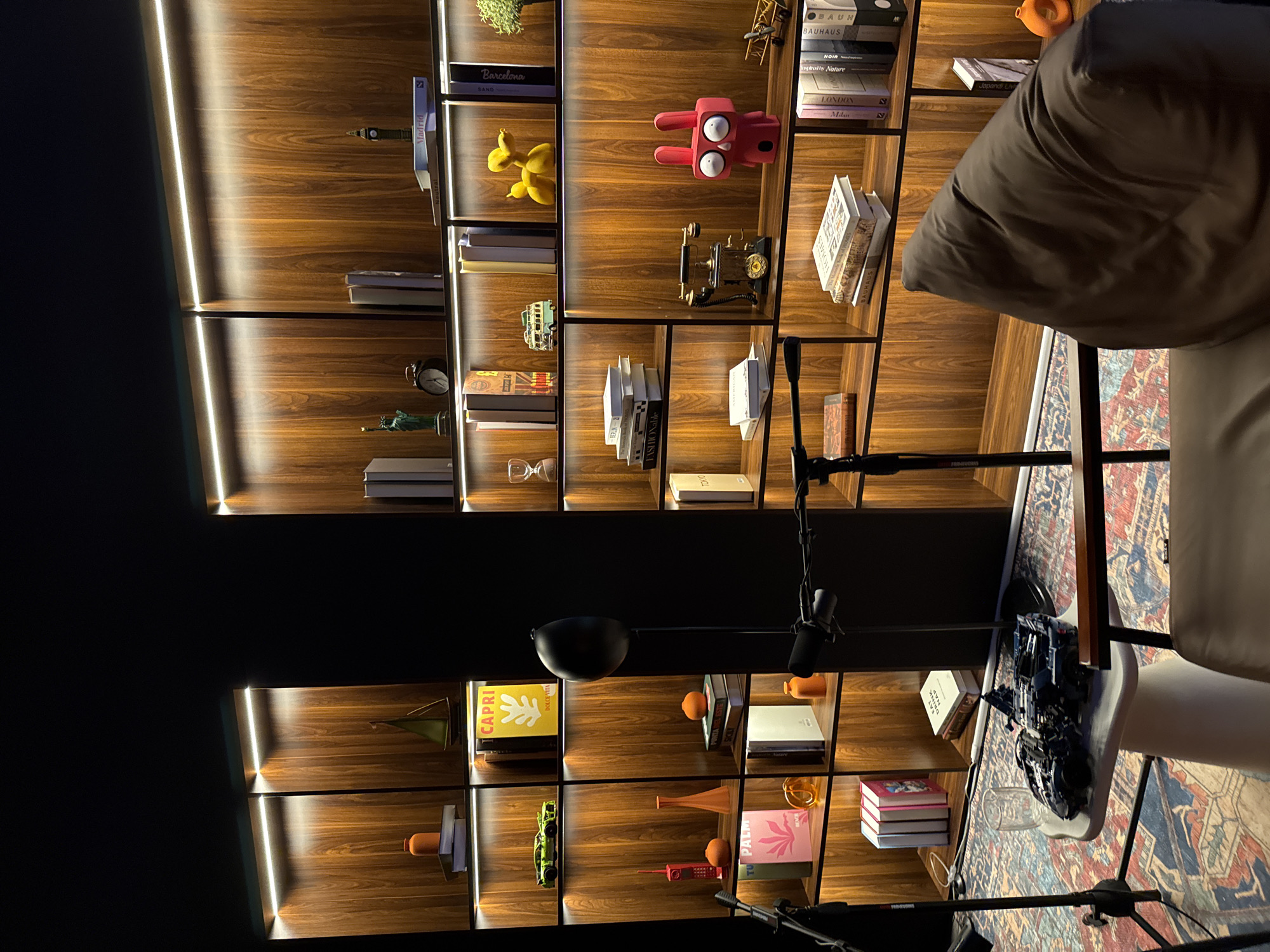 Here is a description of the image:
The image displays a floor-to-ceiling bookshelf unit with wooden shelves and integrated LED lighting along the vertical edges. The shelves contain an assortment of decorative objects, books, and figurines. Some visible titles on the books include "Barcelona SAND", "Madrid", "BAUN", "BAUHAUS", "NOIR", "Londin Milan", "CAPRI DOLCEVITA", "TU PALM", "Japandi Livi", and "Tokyo."
In the foreground, a person sitting in a chair is partially visible in the right. A microphone and related equipment such as stand are also in view, indicating it might be a recording or broadcast setup. A patterned rug can be seen underneath the chair and equipment.
The image does not reveal any definitive information regarding the location as the city of Warsaw, Poland.