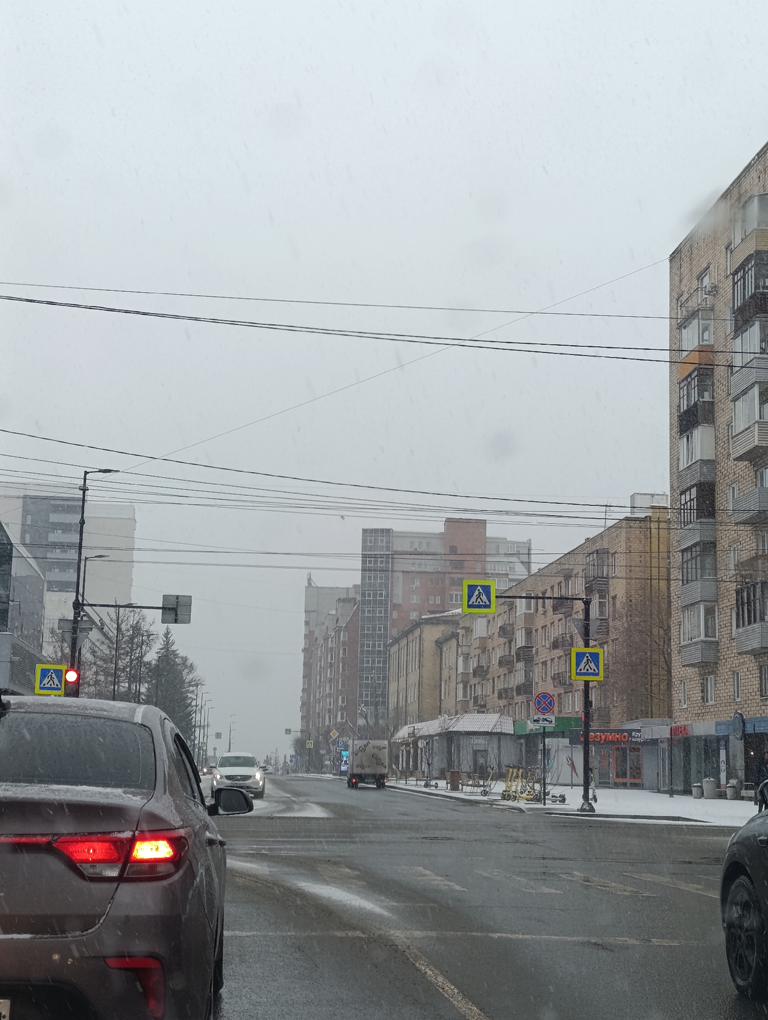 The image depicts a snowy, overcast day on a city street in Krasnoyarsk, Russia, viewed from inside a vehicle. In the immediate foreground on the left, the rear of a dark brown or reddish sedan is prominent, with its red taillights illuminated, indicating it is stopped at a red traffic light. Snowflakes are visible on the car's surface and in the air. A partial view of a black car is seen on the far right.
The street ahead is wet and slushy with patches of snow and tire tracks. A white SUV and a white truck are visible in the mid-distance, moving away from the viewer. The road is flanked by multi-story buildings, mostly constructed from brick, showcasing a mix of architectural styles common in urban Russian settings, with numerous balconies. Overhead power lines span across the muted grey sky.
Several yellow pedestrian crossing signs, featuring a black walking figure, are mounted on poles along the street. A red traffic light is visible on the left. Other street signage includes a 'no stopping or parking' sign (red circle with a diagonal line on a blue background). On the sidewalk near the buildings, a group of yellow electric scooters is parked.
Visible Cyrillic text on storefronts includes "РЭУМНО", "Кэш" (meaning Cash), and possibly "СНЭК" (Snack) on various commercial establishments. The overall scene conveys a cold, wintery atmosphere with ongoing snowfall, contributing to the dull daytime lighting.