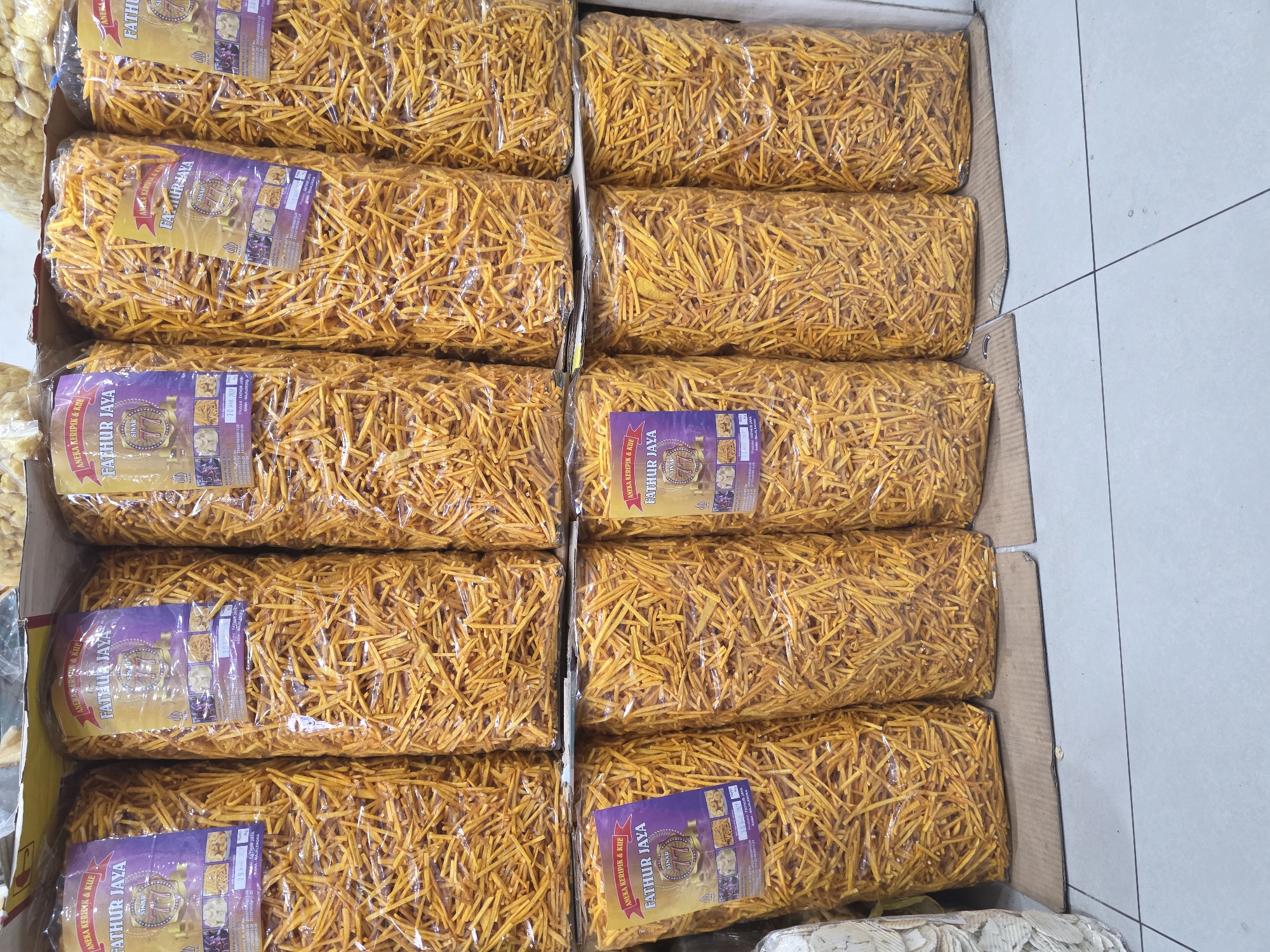 The image displays a retail setting, likely a shop or market stall in Mekarsari, Indonesia, stocked with large, clear plastic bags filled with a crunchy, golden-yellow snack resembling fried potato sticks. The snacks are arranged in multiple rows on shelves or tiered displays. Each bag is branded with a purple and yellow label featuring the text "ANEKA KERIPK & KUE FATHUR JAYA SINAR 77" and various small images of food items. The overall impression is one of abundance and product display.