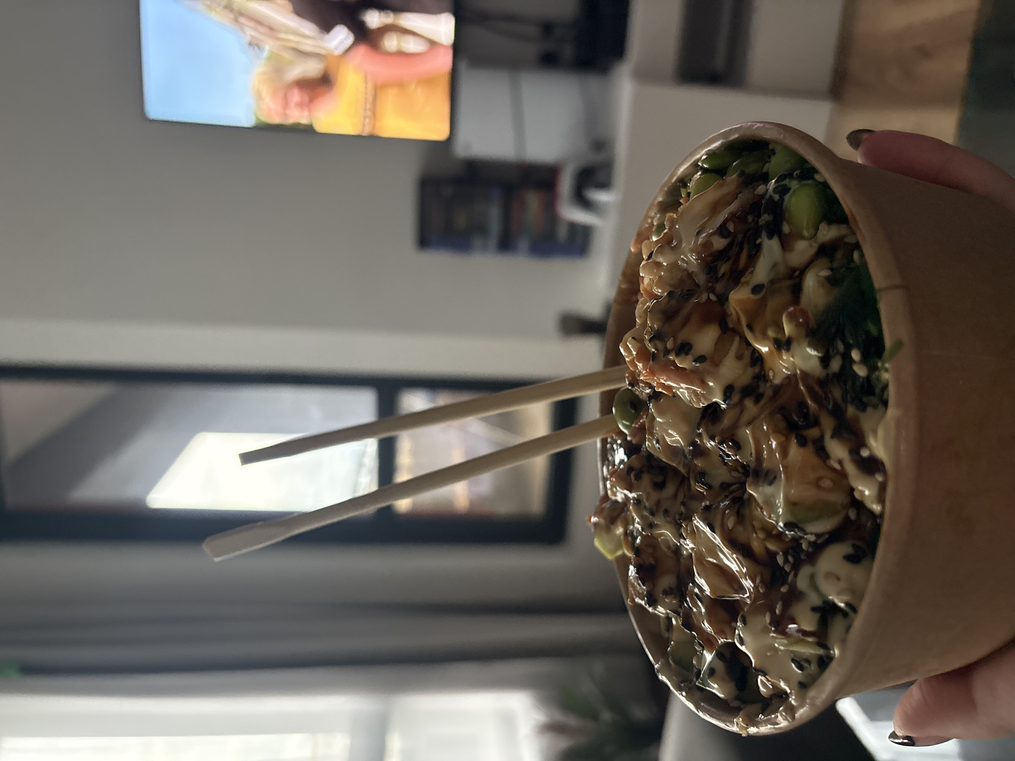 An overhead, slightly angled shot captures a person holding a large brown paper bowl filled with a meal, likely a poke bowl, with two wooden chopsticks inserted. The hand, visible on the right, has dark nail polish. The food within the bowl is covered in a mixture of creamy white and glossy dark sauces, and features green edamame beans, white and black sesame seeds, and what appears to be protein and other vegetables.
The background is softly blurred, indicating a shallow depth of field that keeps the food in sharp focus. In the upper left quadrant of the background, a television screen is visible, displaying a brightly colored image of a person with blonde hair and an orange-yellow garment. Below the TV, there are indistinct shelves or a cabinet holding various blurry objects. The lower background shows a bright area, possibly a window, suggesting an indoor setting like a home living room or dining area. The overall lighting is soft and ambient. No text is visible in the image.