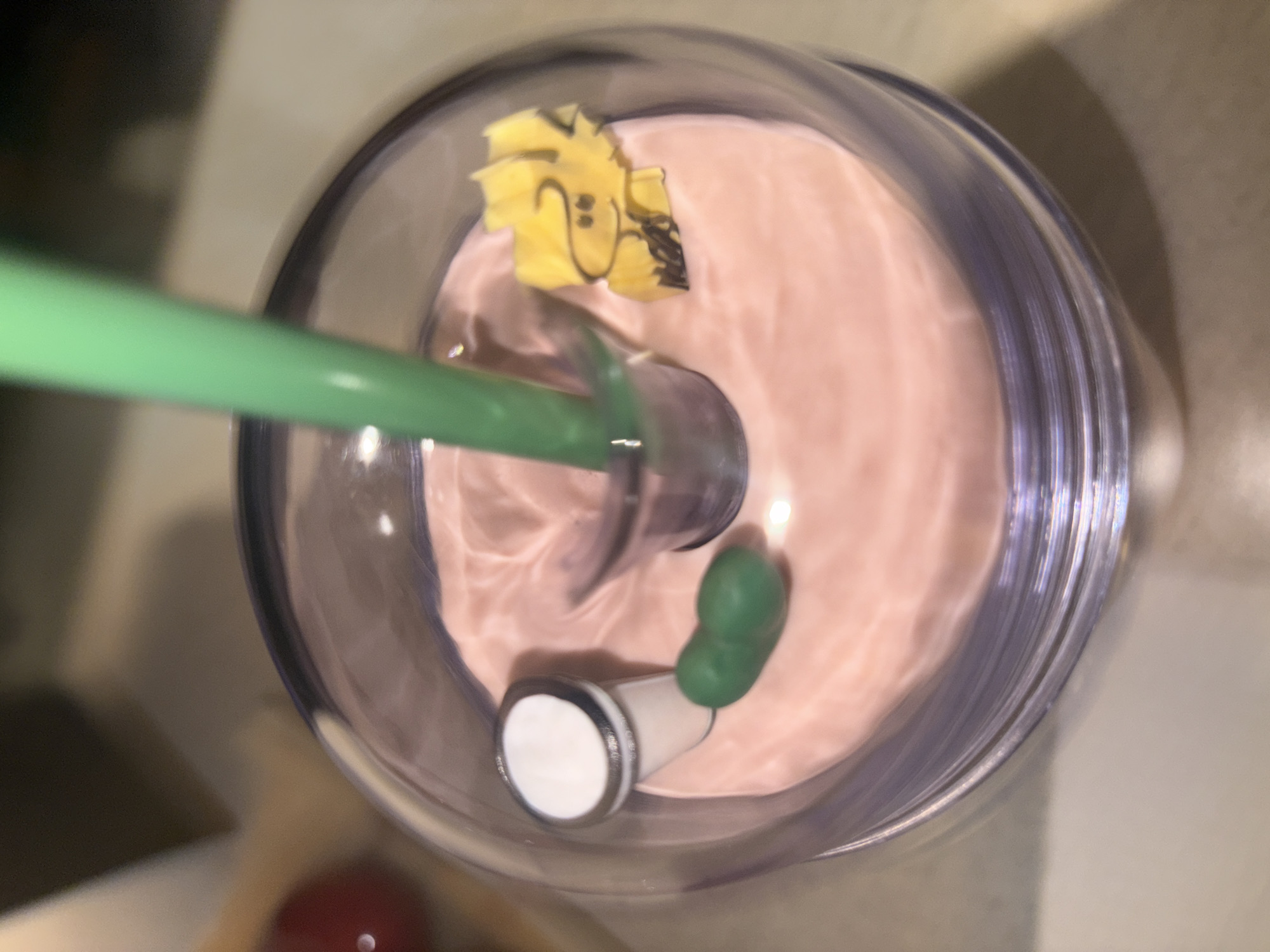 A slightly blurred, overhead shot depicts a translucent tumbler filled with a pink, swirled liquid. A green straw is inserted into the center. A yellow, cartoon-like decoration rests on the liquid's surface. A white and gray stopper, topped with a green object, is also present. The tumbler sits on a neutral-toned surface. This photo was taken in Puebla, Mexico.