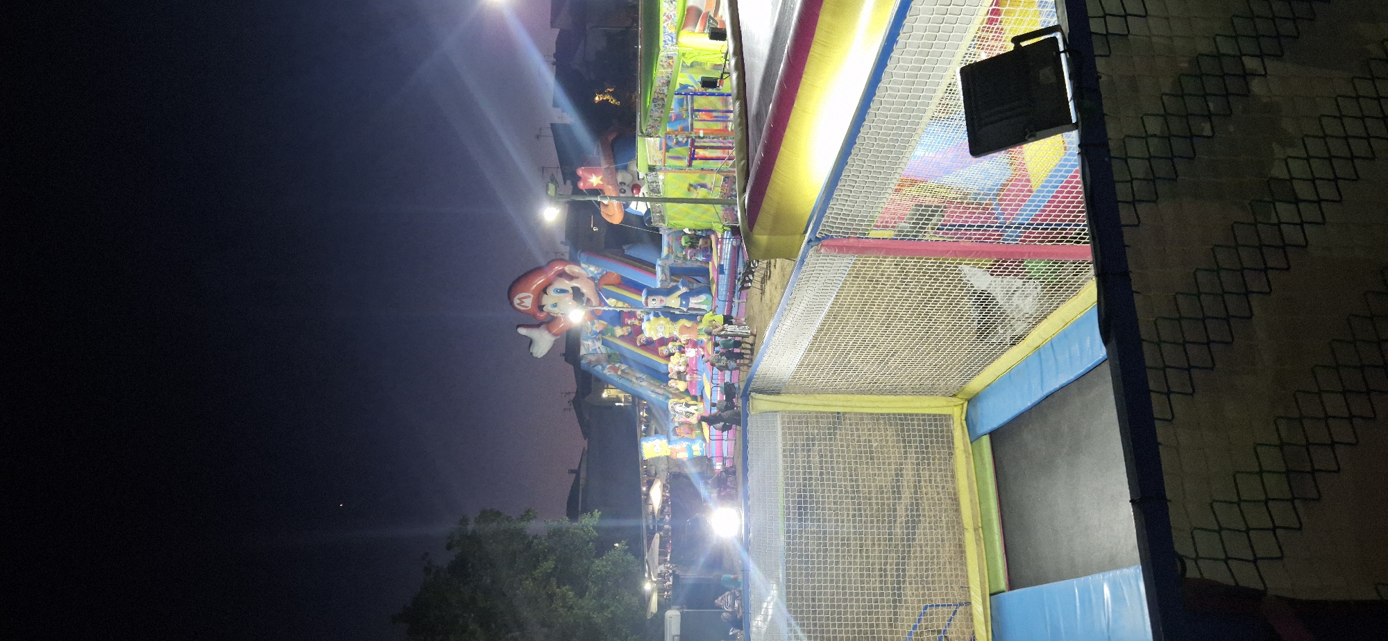 The image depicts a nighttime scene at an outdoor amusement area in Navaluenga, Spain, likely a fairground or park. In the foreground, a trampoline with blue padding and a netted enclosure is visible. Beyond the trampoline, a large inflatable structure dominates the scene, featuring a prominent Mario character at the top of a slide. Various other cartoon characters, including some from The Simpsons, are also integrated into the inflatable. Several people are present around the inflatable structures, some appearing to be children and adults enjoying the attractions. Bright artificial lights illuminate the area, creating a vibrant and festive atmosphere against the dark night sky. In the distance, buildings and trees are dimly silhouetted.