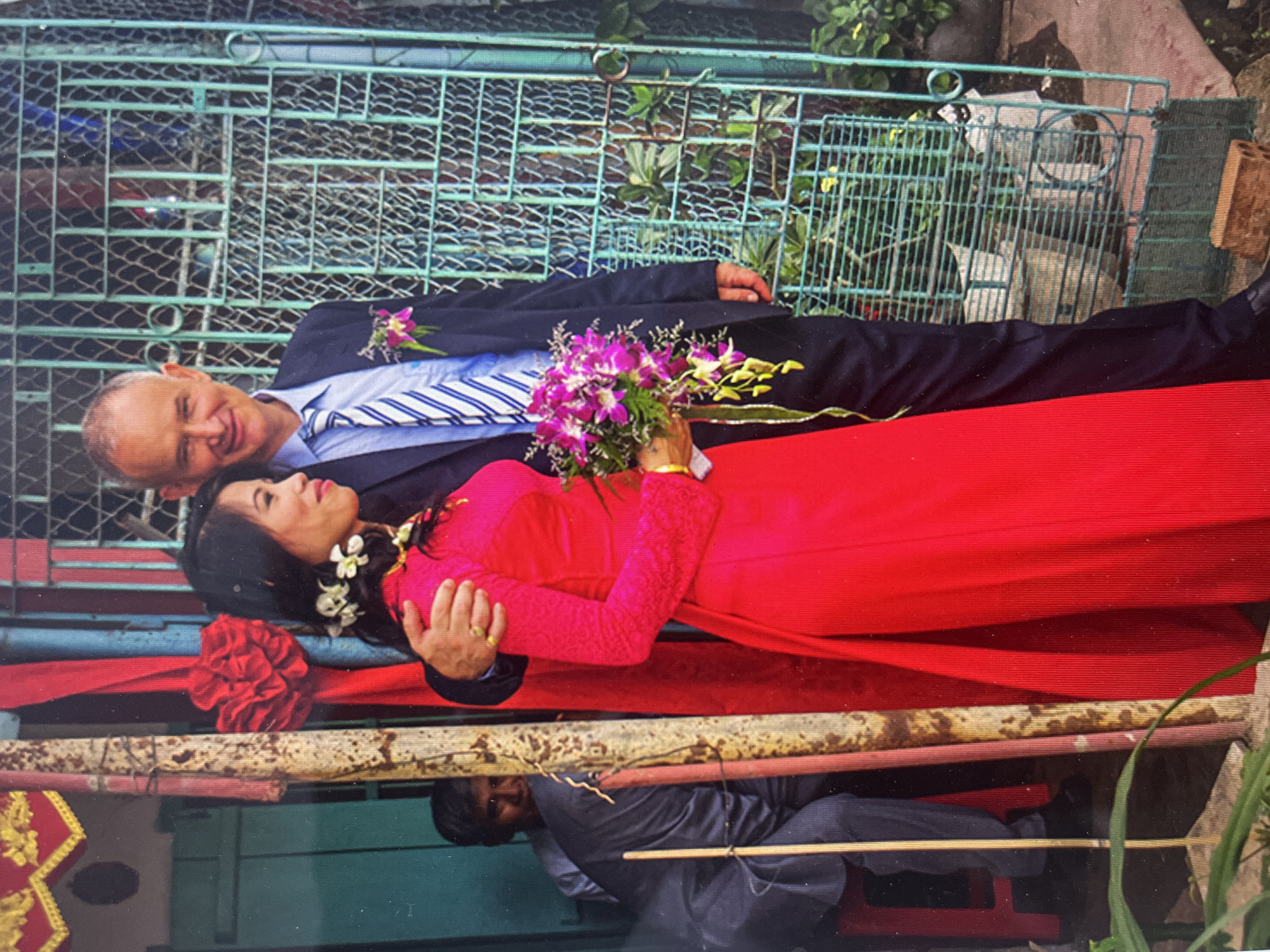 A photograph depicts a wedding scene, likely taken in Bac Nha Trang Ward, Vietnam. A Caucasian man in a navy suit, light blue dress shirt, and blue and white striped tie stands with a woman in a bright red Áo dài, the traditional Vietnamese dress. The woman holds a bouquet of purple orchids and yellow flowers. They are standing on or near a bamboo and red fabric-covered structure. Beneath the structure, a person is seated. The background includes a weathered green metal fence and some vegetation.