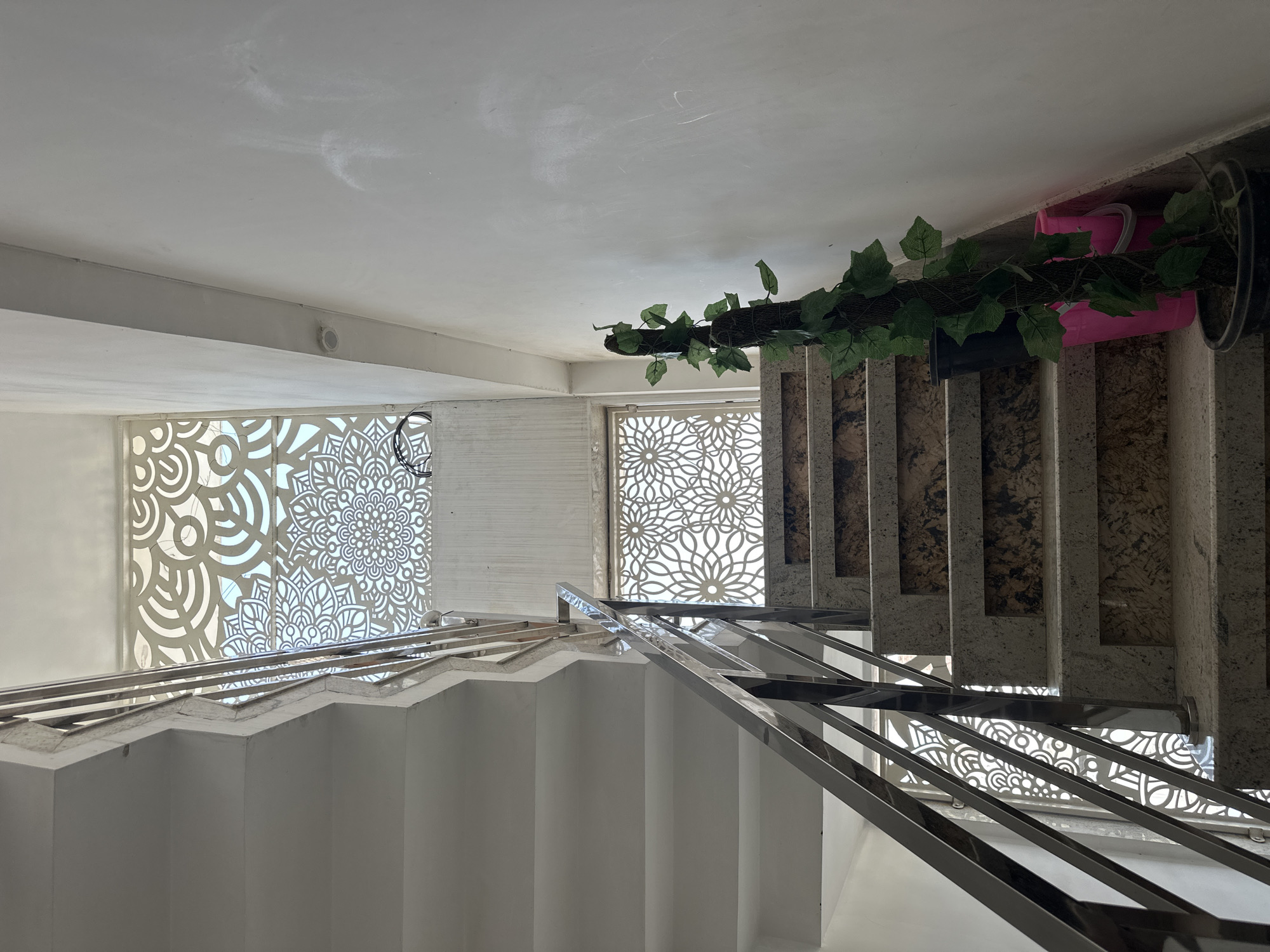 The image depicts the interior of a building, likely in Vrindavan, India, based on the architectural details. The scene focuses on a staircase with white steps and stainless steel railings. The stairwell has decorative window panels featuring mandala-like patterns. Artificial vines and plant pots are placed along the staircase. There are no people visible in the frame. The environment appears well-lit and modern.