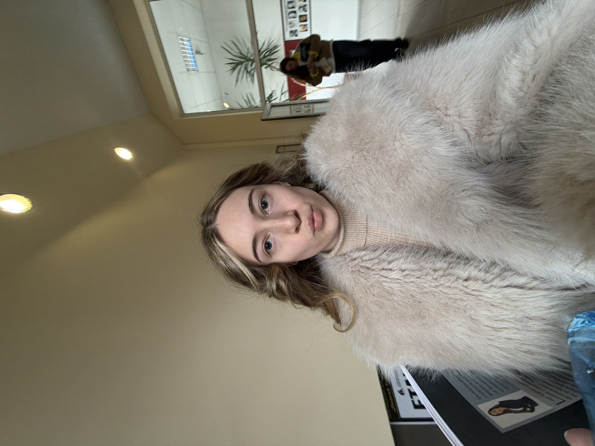 A young woman with blonde hair is wearing a large, light-colored fur coat and a beige turtleneck sweater. She appears to be indoors, potentially in a lobby or waiting area, as evidenced by the blurred background which includes a large window with a view to the outside, other people, and potted plants. The setting resembles an office in the city of Plovdiv, Bulgaria. The lighting is bright and even, suggesting artificial illumination. In the foreground, there is a partially visible object, possibly a magazine or a document. The woman is looking directly at the camera.