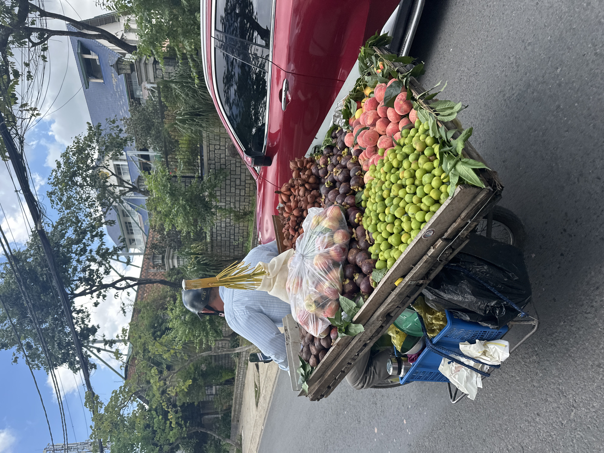 A man wearing a light blue striped shirt and a black helmet rides a motorcycle on an asphalt road in Lâm Viên - Đà Lạt, Vietnam. The motorcycle is equipped with a wooden cart filled with various fruits. The cart contains an abundance of small green fruits, likely plums or limes, a pile of mangosteen, a large plastic bag filled with red and pink fruits, and a cluster of salak fruits. To the left of the cart, a blue plastic basket holds a metal thermos and a green plastic bowl, along with other items. Behind the motorcycle, a red car is parked. The background features trees, a brick wall, a fence, and a building with a blue tiled roof. The sky is cloudy.