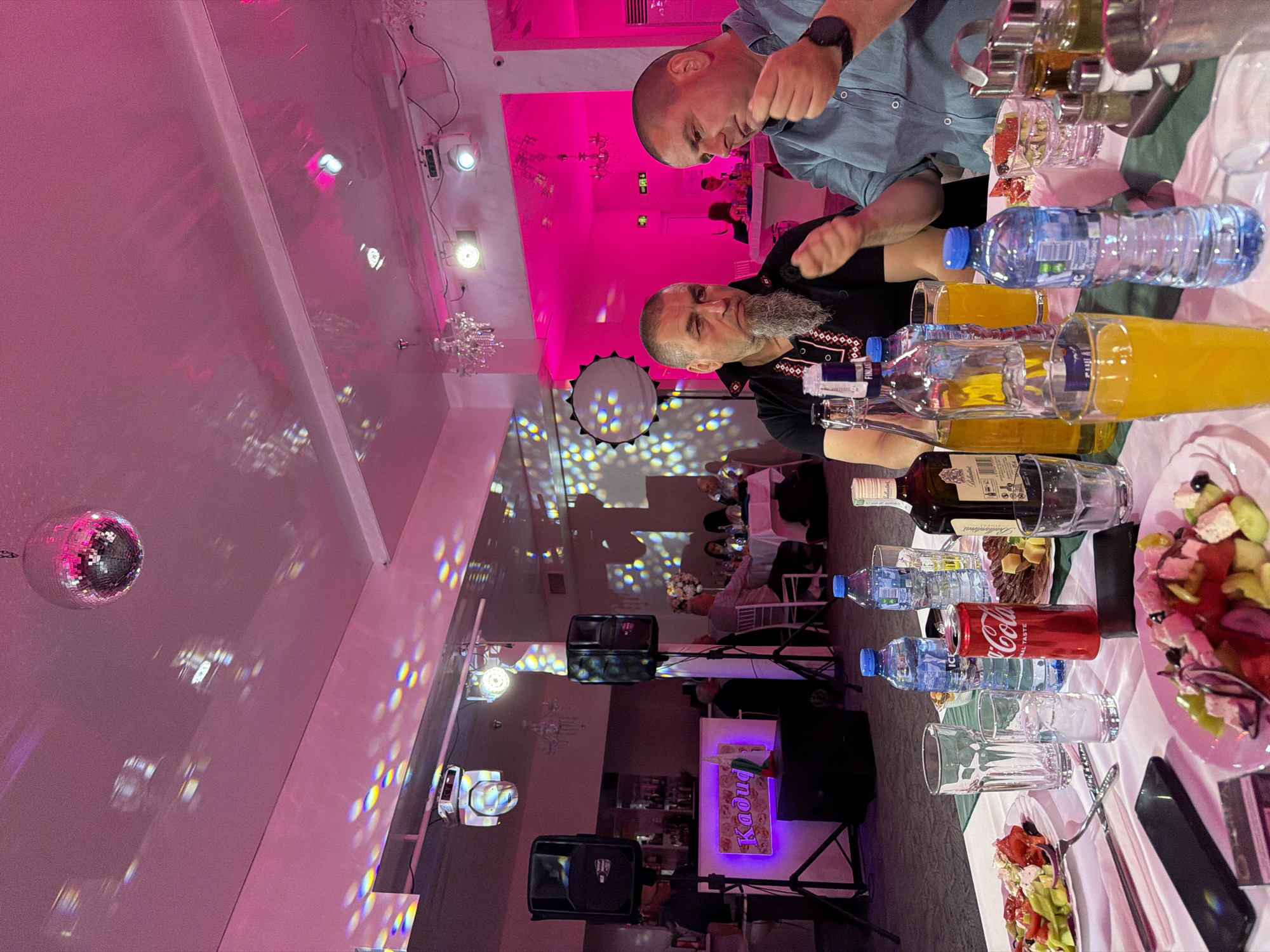 The image appears to be taken inside a function hall, likely in Plovdiv, Bulgaria, given the text "Кадир" visible on a lit sign. The hall is lit with pink and purple lighting, and there are disco balls and spotlights on the ceiling, suggesting a celebratory event.
Two men are prominent in the foreground, partially obscuring a table laden with food, drinks, and glassware. The man on the right is wearing a light blue shirt and a watch. The other man is wearing a black patterned shirt and has a grey beard. Both men are turned towards the camera and appear to be engaged in a conversation or reacting to something.
The table features several bottles of water, Coca-Cola, and alcohol, along with glasses and plates of salad. In the background, other guests are seated at tables. There is a sound system to the left of the image, indicating that the event may involve music or entertainment. Overall, the image captures a social gathering or celebration.