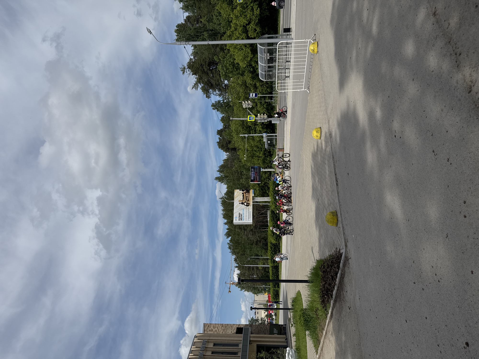 This photograph captures a daytime street scene, likely in Perm, Russia, during what appears to be a cycling event or training session. The perspective is from a sidewalk or road shoulder, looking down a street lined with trees and urban infrastructure.
In the midground, a group of cyclists, many wearing helmets and athletic gear, are gathered with their bicycles. They appear to be in a staging area or near a course, suggesting an organized activity. Some white metal barriers are set up along the edge of the road, potentially for crowd control or defining a path for the event. Several yellow, dome-shaped road safety bollards are visible on the asphalt.
Further down the street, there are traffic lights, pedestrian crossing signs, and a bus stop shelter. A street light pole extends into the upper frame. The background is dominated by a dense line of green trees. Above the trees, a large billboard is visible, featuring images of a person on a bicycle and text. The largest discernible text on the billboard reads "МУЗЫКА ЗВУЧИТ" (Music Sounds/Plays) and "АДРЕНАЛИН" (Adrenaline), with a phone number below.
To the left of the scene, part of a modern brick building is visible in the foreground, and further in the distance, a construction crane can be seen against the sky. The sky is bright with a mix of blue patches and scattered white and gray clouds, indicating fair weather. Shadows are cast on the ground, suggesting it is mid-day. The road surface is asphalt, and there are areas of grass and shrubs along the sidewalk.