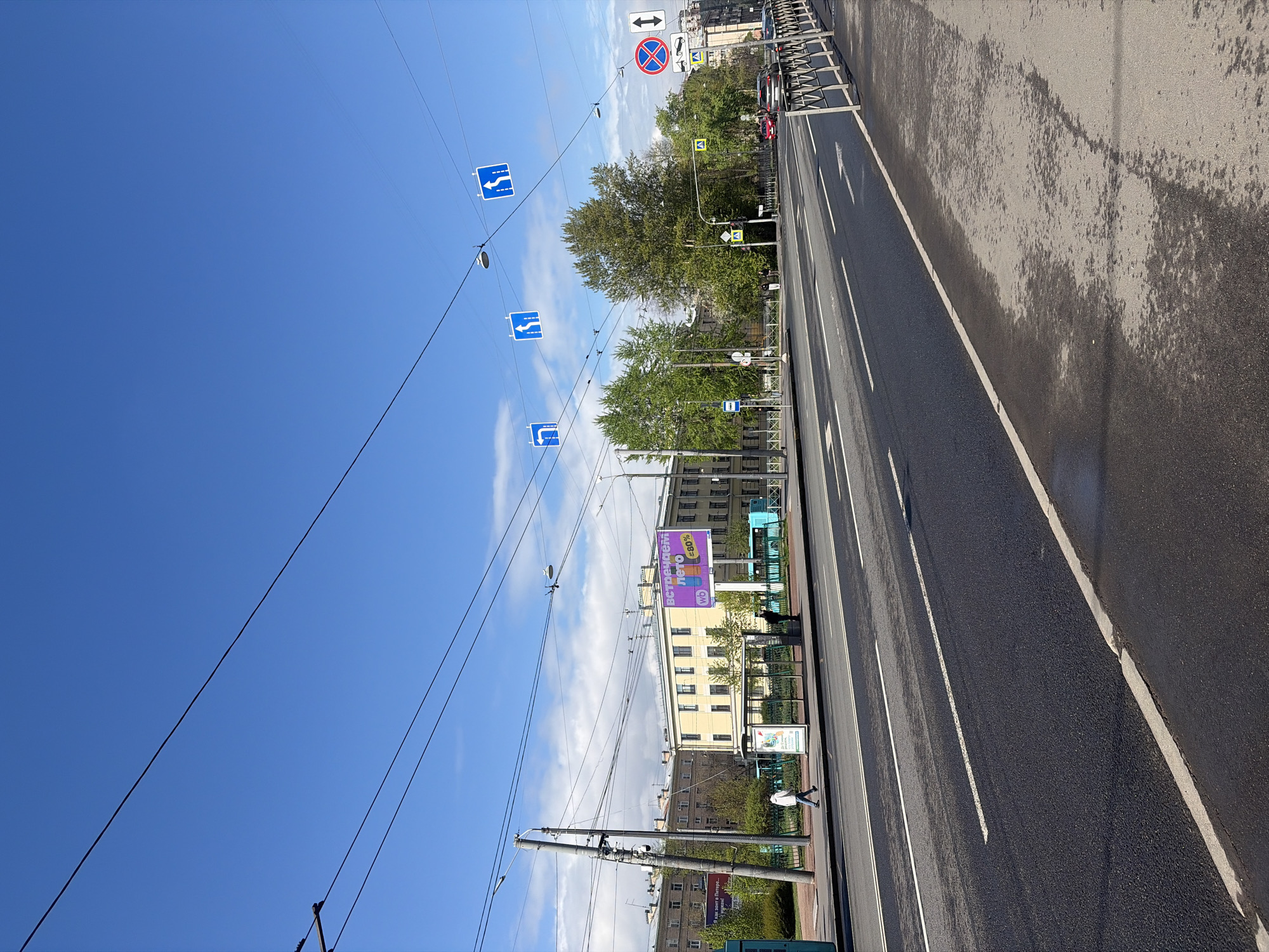 The image captures a street scene in Saint Petersburg, Russia. The view is oriented lengthwise along a multi-lane roadway with clear lane markings. The asphalt shows signs of recent rain or washing, with some areas appearing darker and damp. A sidewalk runs parallel to the roadway, separated by a barrier or a strip of greenery.
Along the sidewalk, there are pedestrian figures, one of whom appears to be walking. Buildings line the street, including a yellow building with multiple windows and a larger, light-colored building with a purple advertising billboard on its side.
Overhead, a network of electrical wires and cables crisscrosses the sky. Various traffic signs are visible, including blue direction signs and a red and blue sign indicating a prohibition of some kind. Trees are interspersed between the buildings and along the street, providing some greenery to the urban environment. The sky is clear with some scattered clouds.