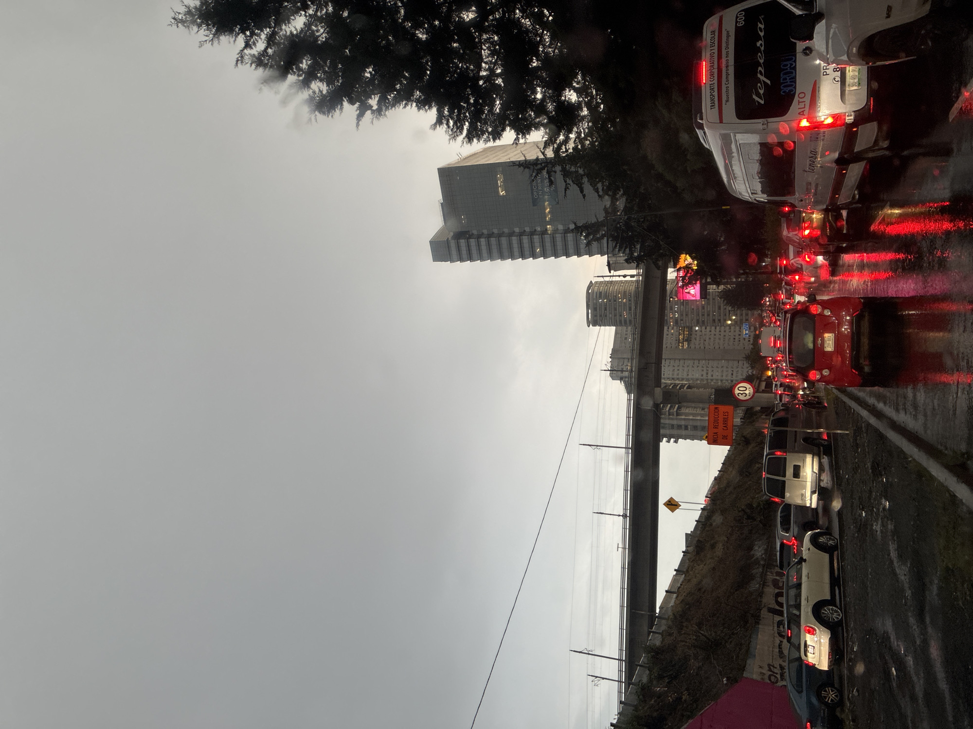 The image depicts a traffic jam on a rainy day in Mexico City, Mexico. A line of cars, vans and trucks are stopped on the wet road. A large office building looms in the background, and a light rail bridge extends across the scene. Road signs indicate lane reduction and a speed limit of 30 km/h. The vans are branded with "tepesa" and "TRANSPORTE CORPORATIVO Y ESCOLAR" indicating a corporate and school transportation service.