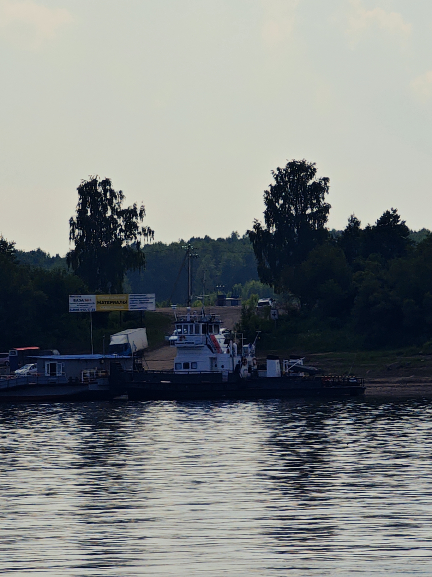 The image shows a wide, calm body of water in the foreground, with ripples reflecting the light. In the midground, a ferry or barge is docked at a shoreline with a sandy or muddy bank. The vessel appears to be a multi-decked structure, with a white cabin on the upper level. There are several vehicles visible on the ferry, including a white car and a red truck. Adjacent to the ferry, on the shore, there are buildings, including a small structure with a blue roof and windows. A large white trailer is also parked nearby. Behind the shoreline, a dense forest of dark green trees creates a backdrop, with two prominent trees with thin branches standing out against the lighter sky. A billboard with Russian text is visible amongst the trees. The sky is overcast and light-colored. The scene is likely a port or a ferry crossing in Erkalovo, Russia, given the presence of Russian text on the billboard and the general landscape.