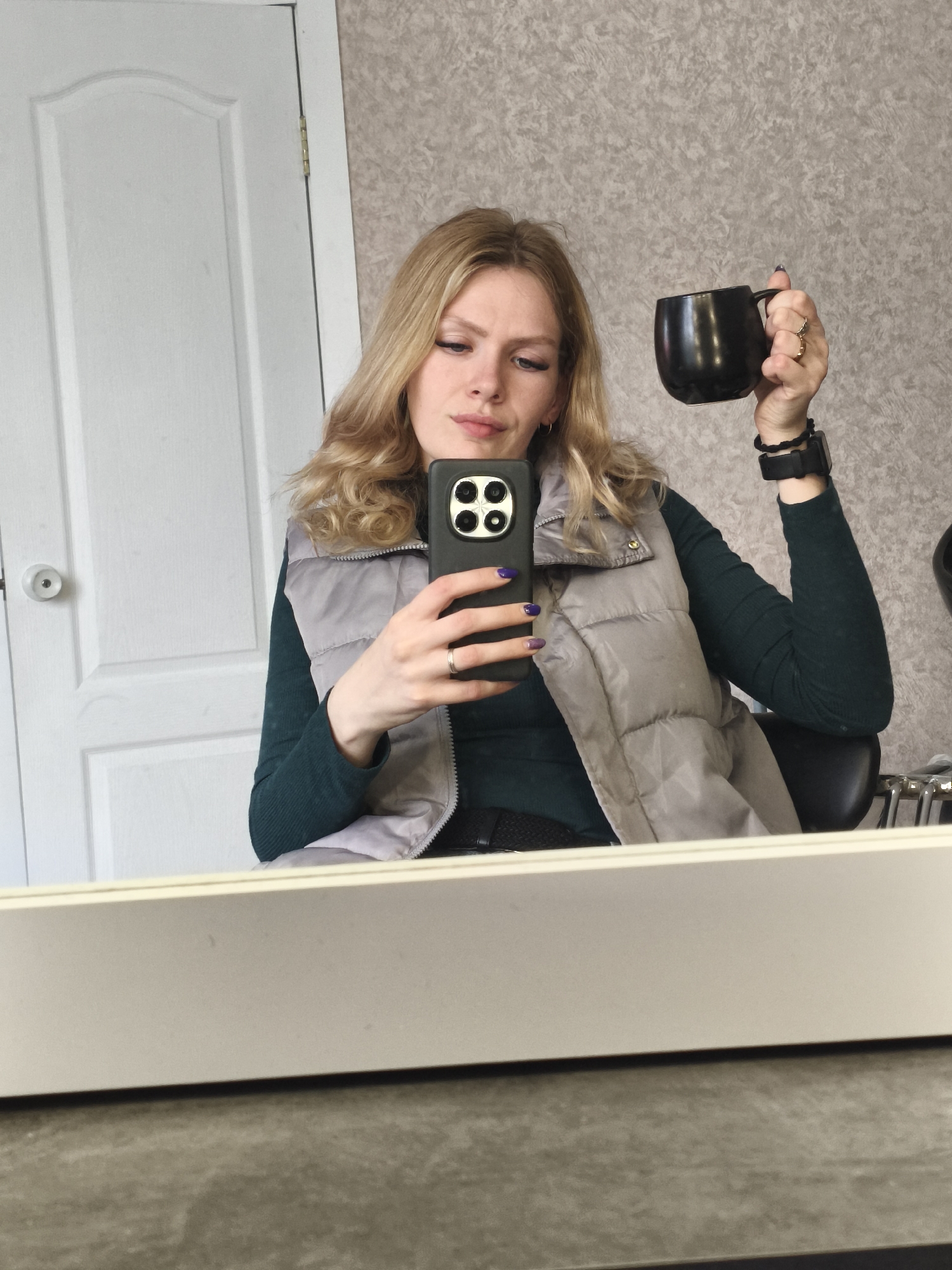 A young adult woman with long, wavy blonde hair takes a mirror selfie in an indoor setting, likely a salon or dressing room in Русский Вожой, Russia. She is reflected from the chest up, looking down at a black smartphone with a quad-camera system, held in her left hand. Her left hand shows purple gradient nail polish and a silver-colored ring. In her right hand, she holds a black mug. A gold-colored ring adorns her right ring finger, and her wrist sports a black smartwatch or fitness tracker, along with a braided bracelet.
She wears a light gray puffer vest over a dark green, long-sleeved ribbed shirt. Her expression is neutral and focused on her phone screen. The background, visible in the mirror, features a textured, light gray wall or wallpaper on the right and a white paneled door with a round doorknob and visible hinges on the left. A portion of a black chair and some metallic objects, possibly salon equipment, are reflected to her right. The mirror itself has a light-colored frame along its bottom edge. The bright, even lighting suggests it is daytime. No visible text is present.