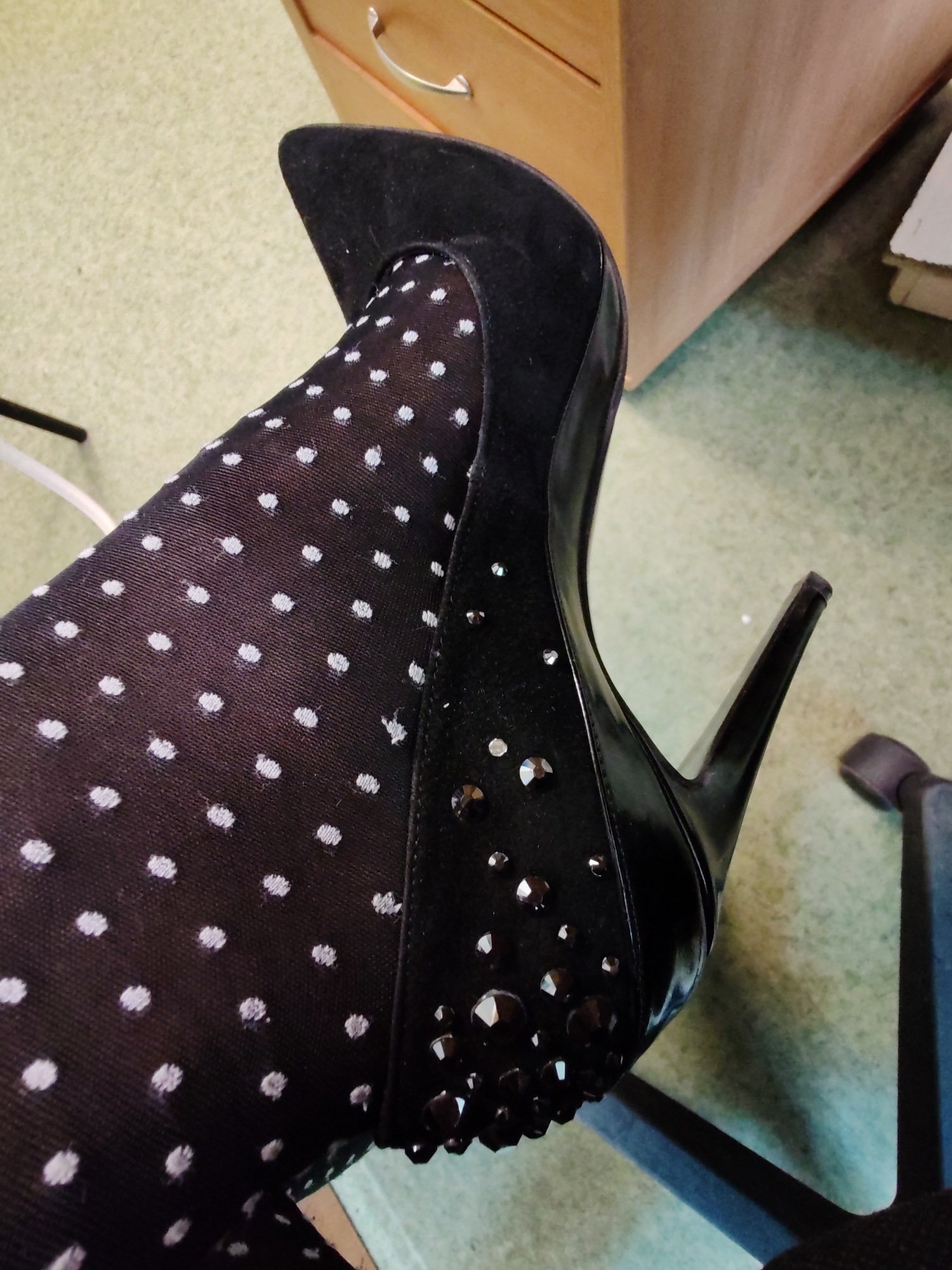 A close-up photograph shows a person's leg clad in black polka dot tights, wearing a black high-heeled shoe. The shoe is adorned with shiny embellishments. The background includes a light-colored wooden cabinet with a silver handle and an office chair. The location is unknown.