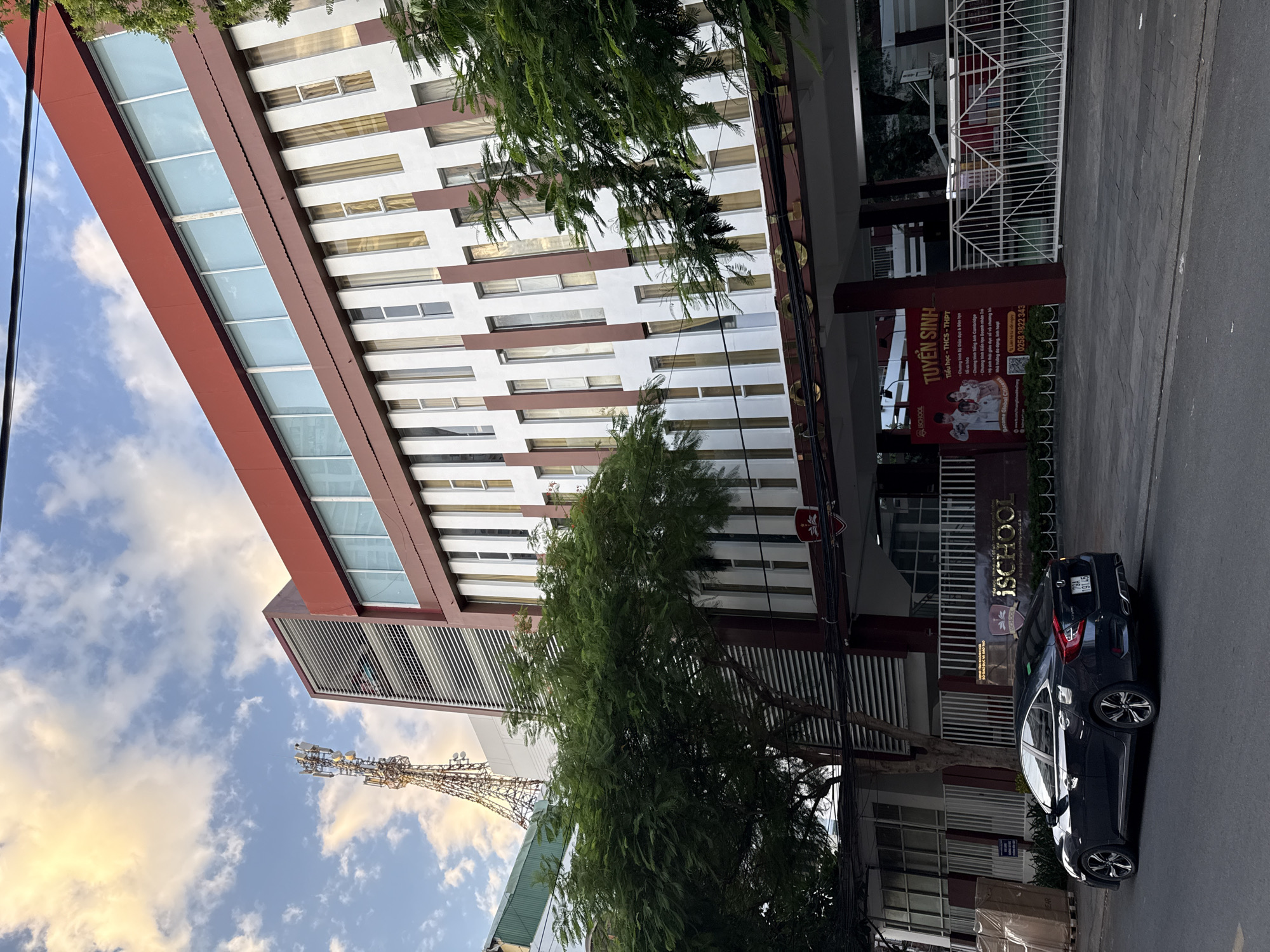 The image depicts the exterior of iSchool Nha Trang, a school located in Nha Trang, Vietnam. The building is modern, with a predominantly red and white facade. A large, multi-story structure with numerous windows and vertical accents dominates the scene. In the foreground, a dark-colored sedan is parked on the street. Lush green trees partially obscure the building and the street. A telecommunications tower is visible in the background against a partly cloudy sky. The entrance to the school features a gate and a large banner advertising student recruitment, written in Vietnamese. The overall impression is one of an urban educational institution in a tropical or subtropical setting.