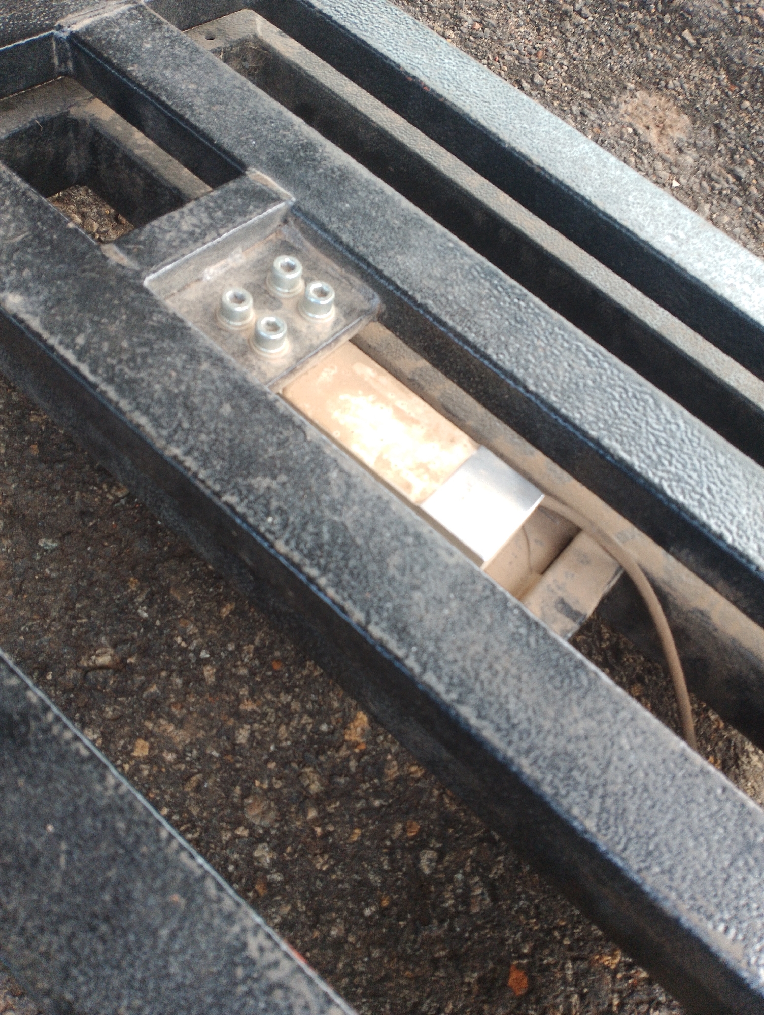 The image shows a close-up, high-angle view of a section of a black metal structure, possibly part of a trailer or a large scale. The structure is composed of thick, rectangular metal bars with a textured surface, likely painted. In the center of the frame, a recessed area reveals a metallic component, possibly a load cell or sensor, with a beige-colored rectangular element and a silver-colored metallic cylinder attached to it. Four silver-colored bolts secure a small metal plate to the structure above this component. A thin, brown cable emerges from the recessed area and curves out of view to the right. The surrounding ground is a rough, dark asphalt or concrete surface with visible small stones and debris. The overall scene appears to be outdoors, with a gritty, industrial aesthetic. There are no people or obvious signs of human activity, and the image's context is limited to the observed objects and environment. The image does not contain enough information to determine its location in Jos, Nigeria.