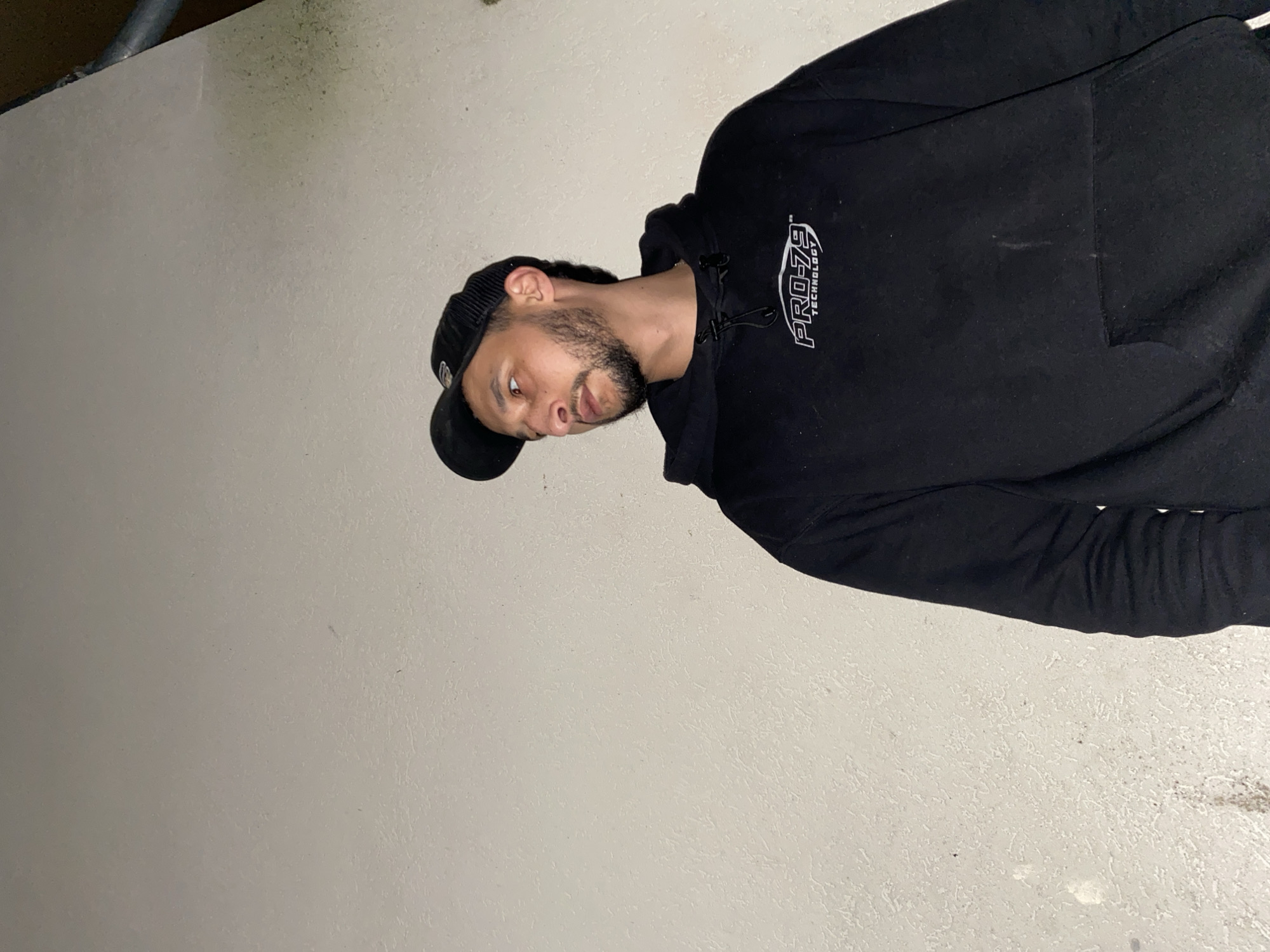 A man with a beard, wearing a black hoodie and baseball cap, is indoors against a gray wall. He is looking to his left and slightly downward. The location cannot be determined from the image.