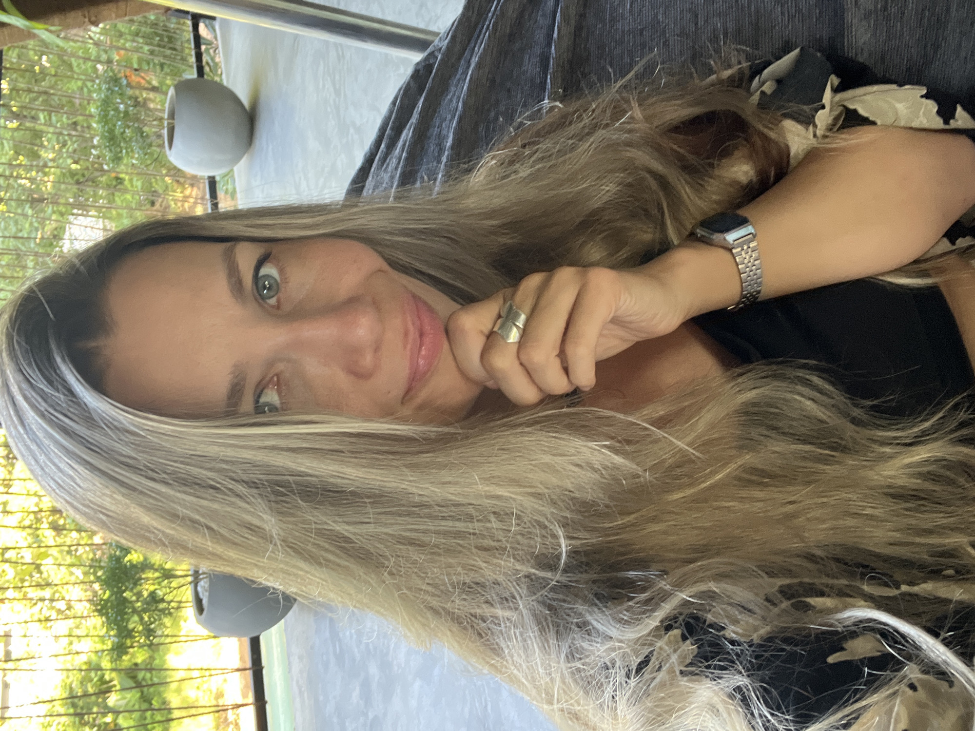 A woman with long, blonde, wavy hair takes a selfie outdoors, resting her chin on her hand. She is wearing a black shirt with a floral pattern on the sleeves and a silver watch on her left wrist. Her eyes are a light green, and she is smiling. Behind her, there is a blurred background of greenery and a gray, rounded planter. The setting appears to be a patio or balcony. The image does not contain information to determine the location within Weligama, Sri Lanka.