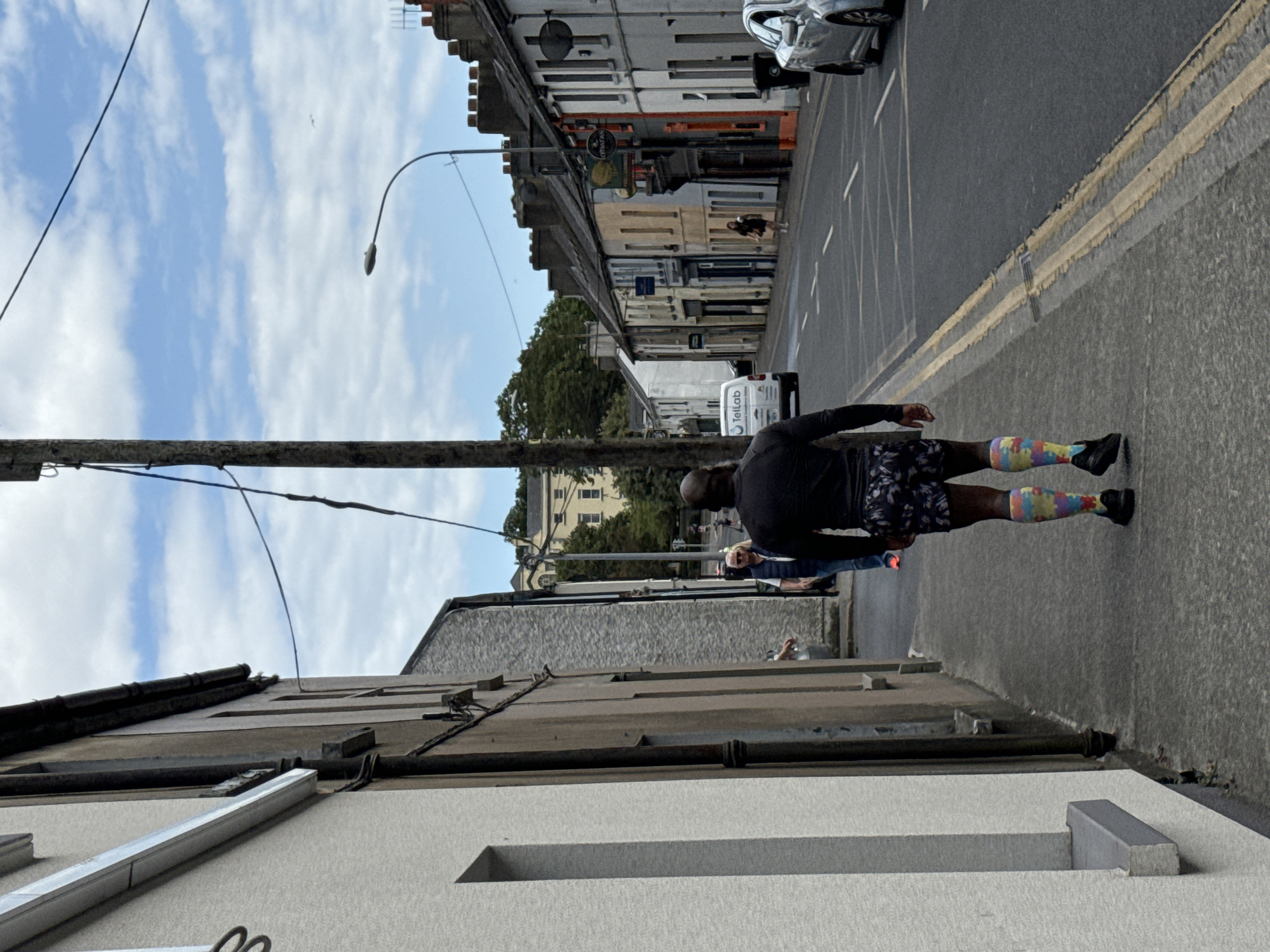 The image depicts a street scene in the city of Waterford, Ireland. In the foreground, a Black man is walking away from the camera, wearing a black long-sleeved shirt, patterned shorts, and colorful compression socks with athletic shoes. He is walking on a sidewalk beside a building. Behind him, further down the sidewalk, a woman in a white vest and blue top is walking. The street itself is paved and has double yellow lines. Several cars are visible on the street, including a silver car on the right side. The buildings lining the street are of varying heights and colors, typical of an urban setting. A white van with the logo "TellLab" is parked on the street. The sky is partly cloudy. The scene appears to be captured during daylight hours.