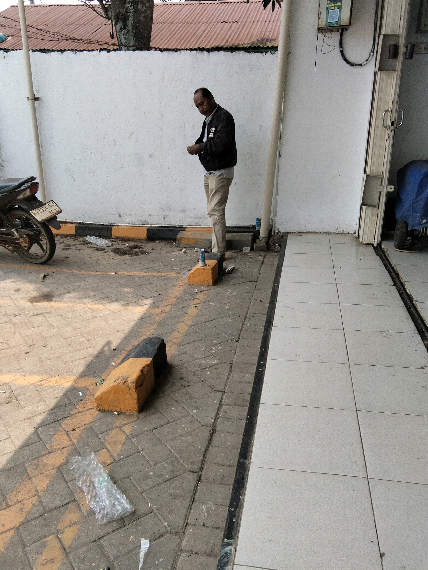 A man, appearing to be in his 30s or 40s, stands near a white wall in what appears to be an outdoor area with tiled and paved surfaces. He is wearing a dark jacket over a light-colored shirt and light-colored pants, and is looking down at his hands. A motorcycle is parked to his left, and several yellow and black concrete barriers are visible on the ground. The scene is littered with debris, including plastic packaging and what appear to be cigarette butts. A metal pole extends vertically along the white wall, and a metal grate runs along the edge of the tiled area. The background shows a corrugated metal roof and some vegetation. The image was taken in Sungai Rebo, Indonesia.
