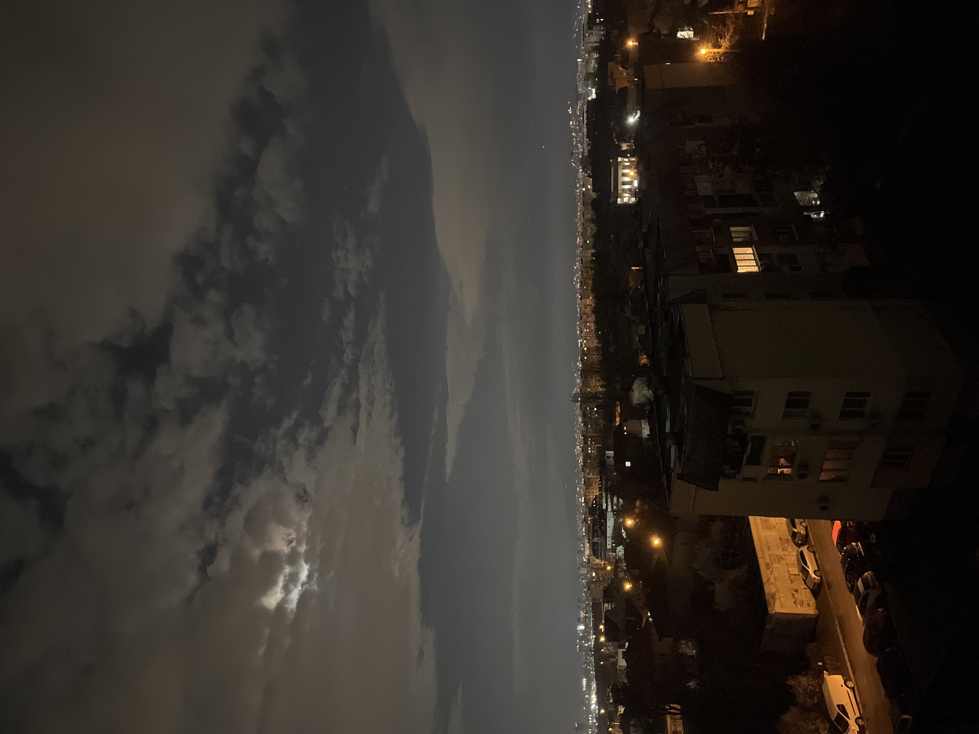 Here is a detailed description of the image:
The image appears to be taken from a high vantage point, possibly a balcony or rooftop, at night. On the left, the night sky is dominated by clouds and the moon is visible. On the right, the cityscape stretches into the distance, with buildings of varying heights illuminated by streetlights and building lights. Along the street, cars are parked. The scene captures the essence of an urban landscape at night, blending natural elements like the night sky with the artificial illumination of the city.
Without further details, the specific location in Sofia, Bulgaria, is difficult to confirm, but the urban density and architectural style suggest a residential area within the city.