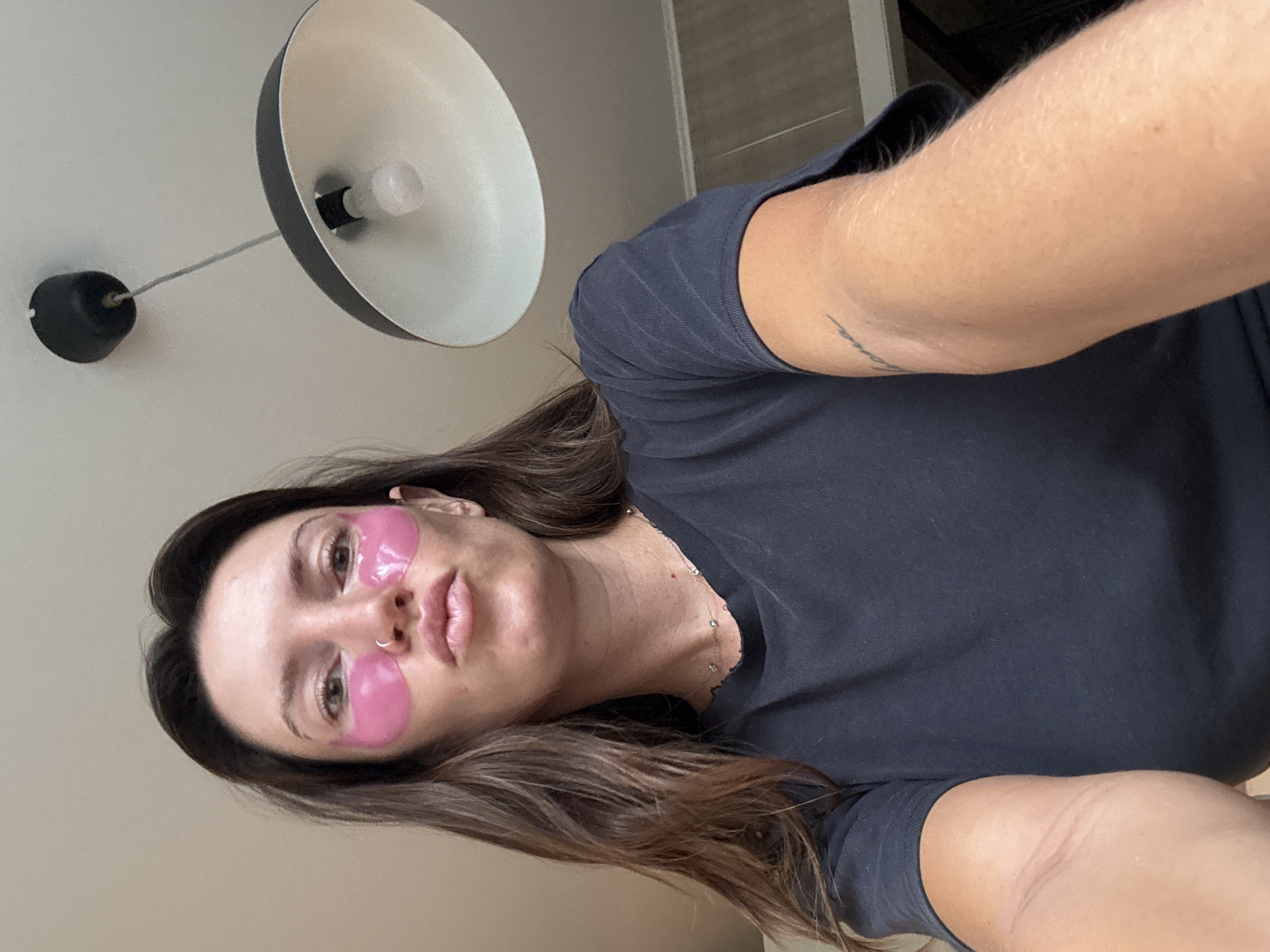 This is a vertical, medium shot selfie of a woman with long, dark brown hair. She is wearing a dark gray t-shirt and has pink under-eye masks on. She is looking directly at the camera. Her expression is neutral. A portion of a ceiling light fixture is visible in the upper left corner of the image. The woman has a small nose ring. A faint tattoo is visible on her upper arm. The overall lighting suggests it is daytime, and the indoor setting appears to be a room with neutral-colored walls. There is no visible text in the image.
