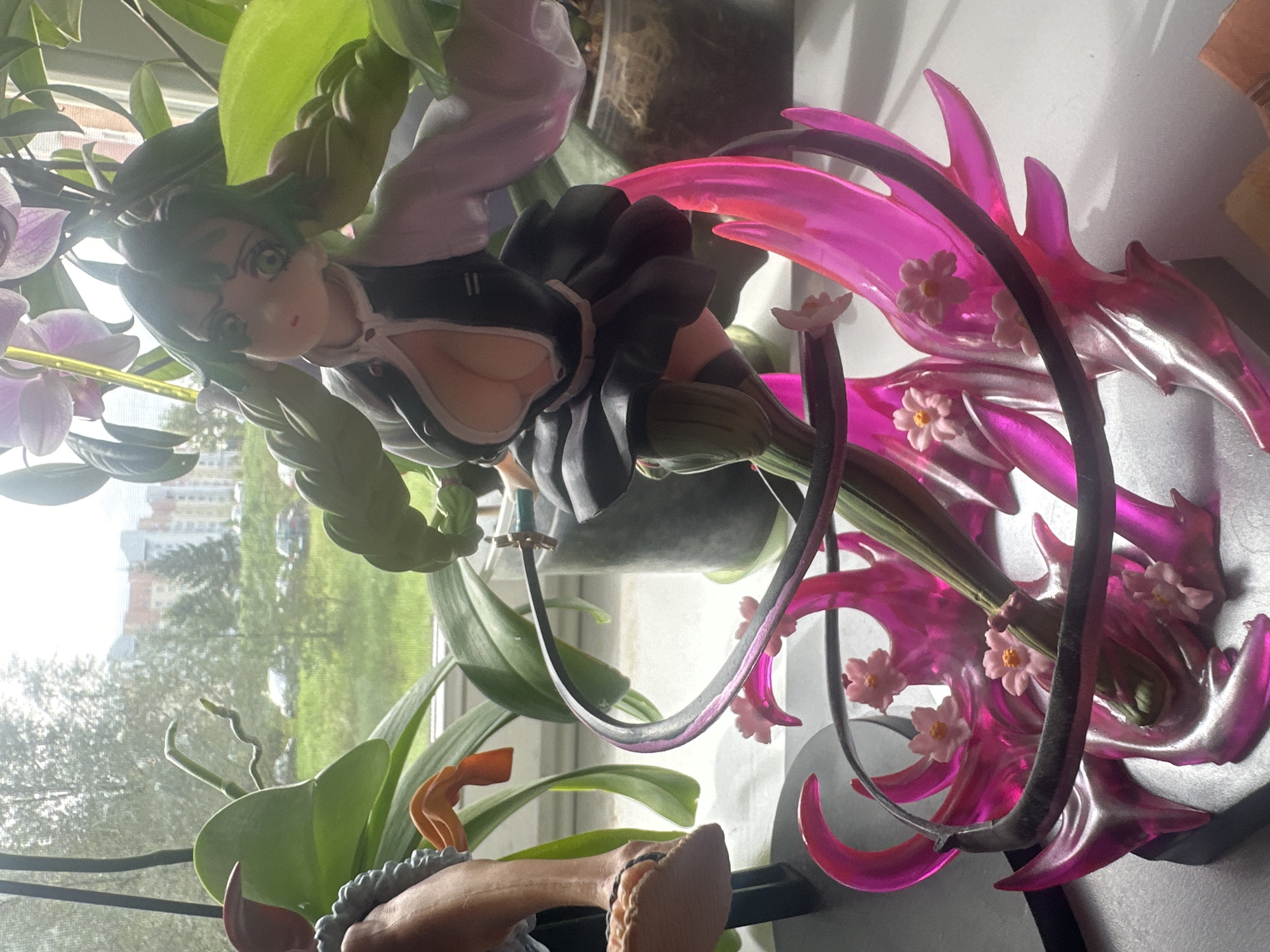 This is a close-up, eye-level, indoor shot of an anime figurine placed on a white surface, likely a windowsill, with a blurred outdoor background. The figurine depicts a young female character with green hair, large green eyes, and a revealing outfit. She is posed dynamically, with her arms outstretched and her body twisted. Her outfit consists of a white shirt with a black vest, a black skirt, and thigh-high stockings. The figurine is part of a larger display, with pink translucent, flame-like structures and small pink flowers forming the base. A dark grey ribbon or band wraps around the base and the figurine. In the background, through a window, a residential area with multi-story buildings, trees, and parked cars is visible, suggesting an urban or suburban environment. The lighting suggests it is daytime, with sunlight streaming in. The specific location in the city of Maladzyechna, Belarus cannot be determined from the image alone.
